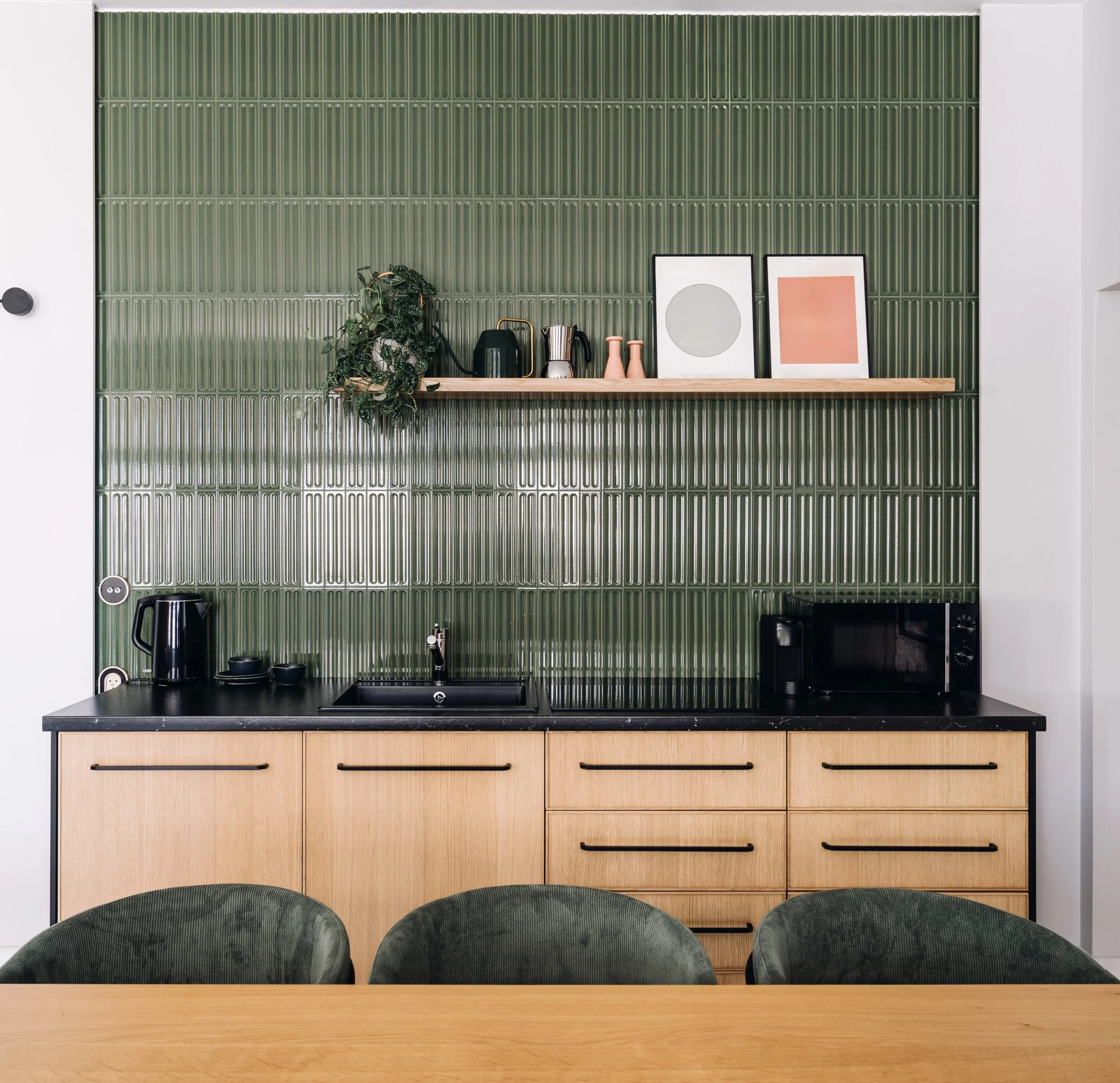 Kitchen with green tiled backsplash, wooden cabinets, and black countertop with a black kettle, framed art, potted plant, and kitchen appliances.