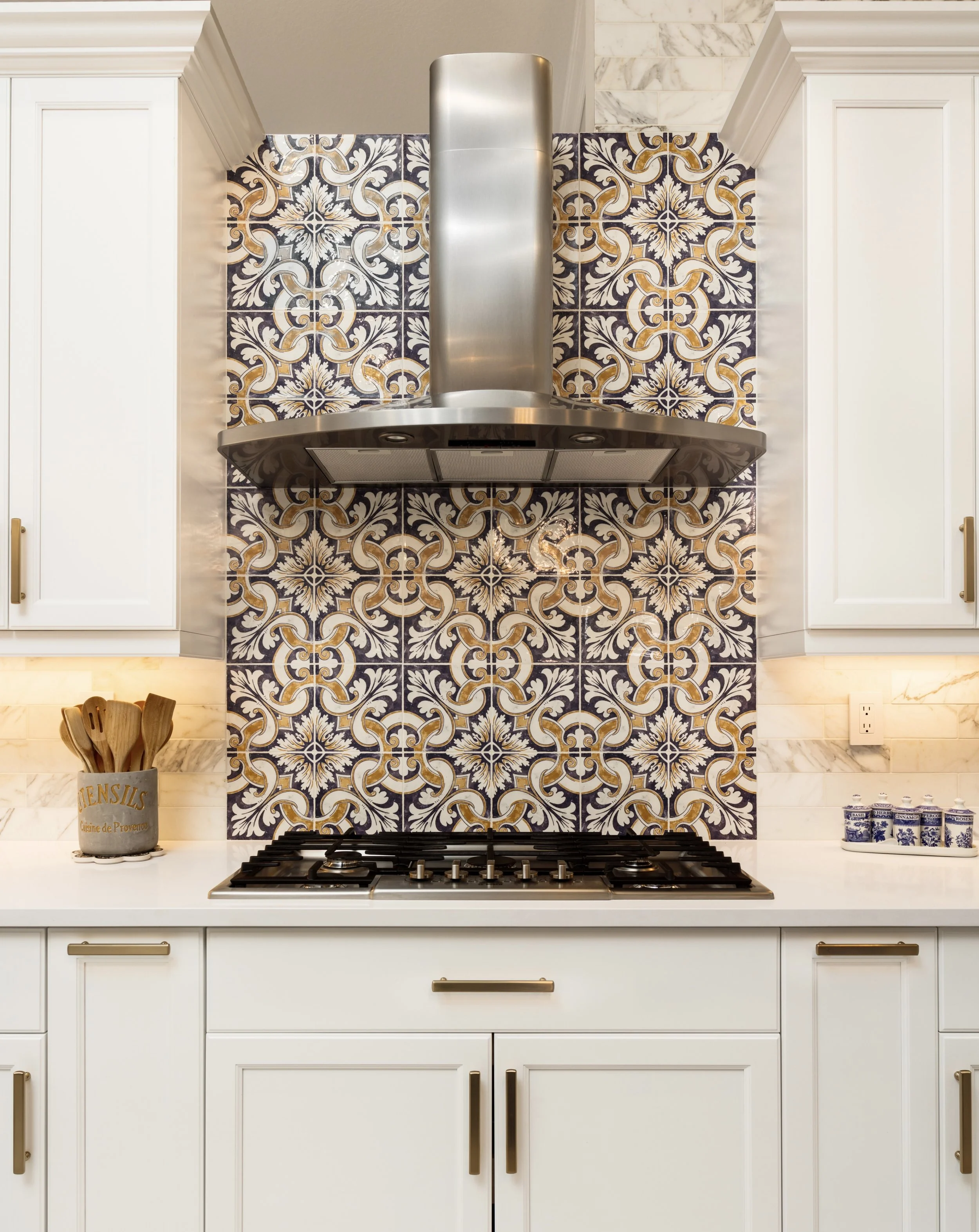 White kitchen with a gas stove, ornate patterned tiles with floral and geometric motifs in navy blue, white, and gold, white cabinetry with gold handles, a pot holding wooden utensils labeled "Ustensils," and a decorative set of blue and white jars, 
