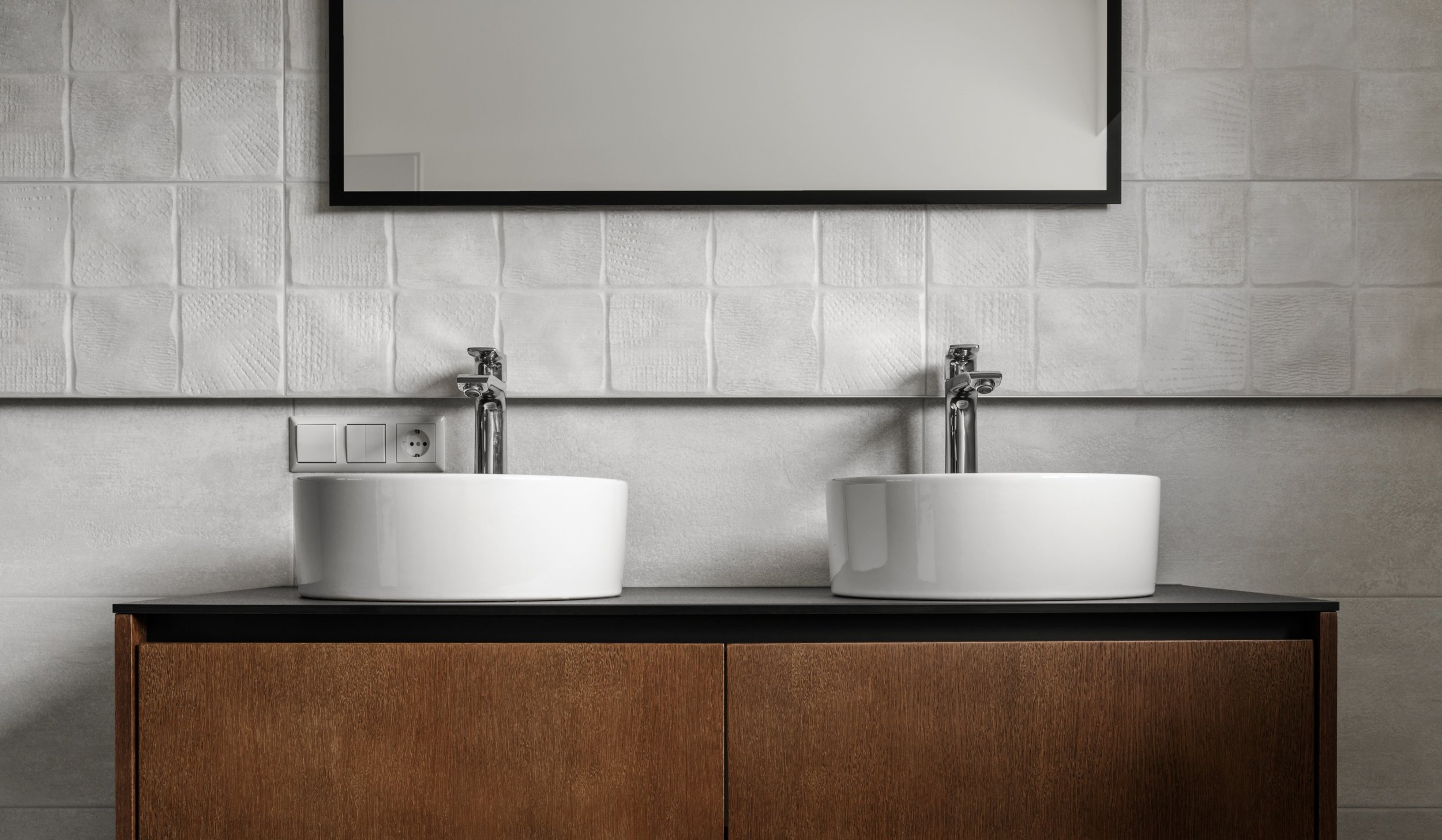 Two modern white sinks with chrome faucets on a black countertop, wooden cabinet below, and a large mirror on a textured white tiled wall.