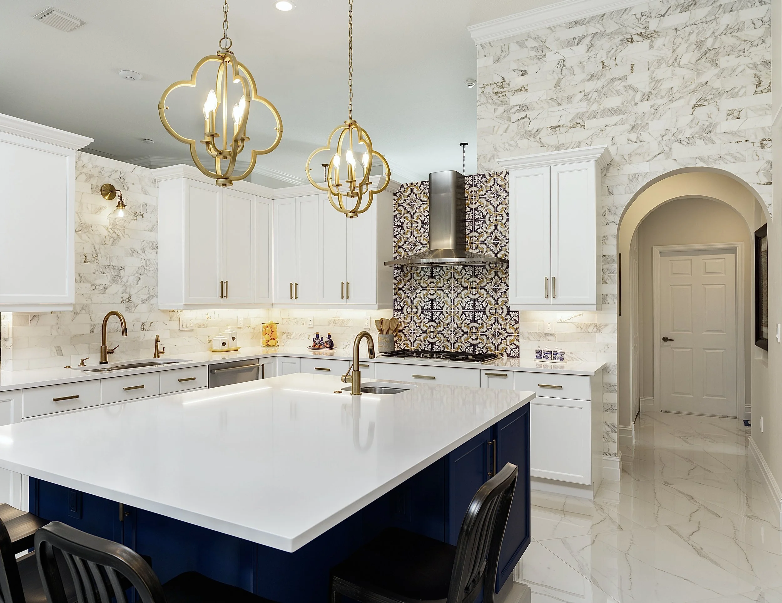 Modern kitchen with white marble backsplash and countertops, blue lower cabinets, white upper cabinets, stainless steel range hood, decorative tile behind stove, gold fixtures, and black chairs around an island with a white countertop.