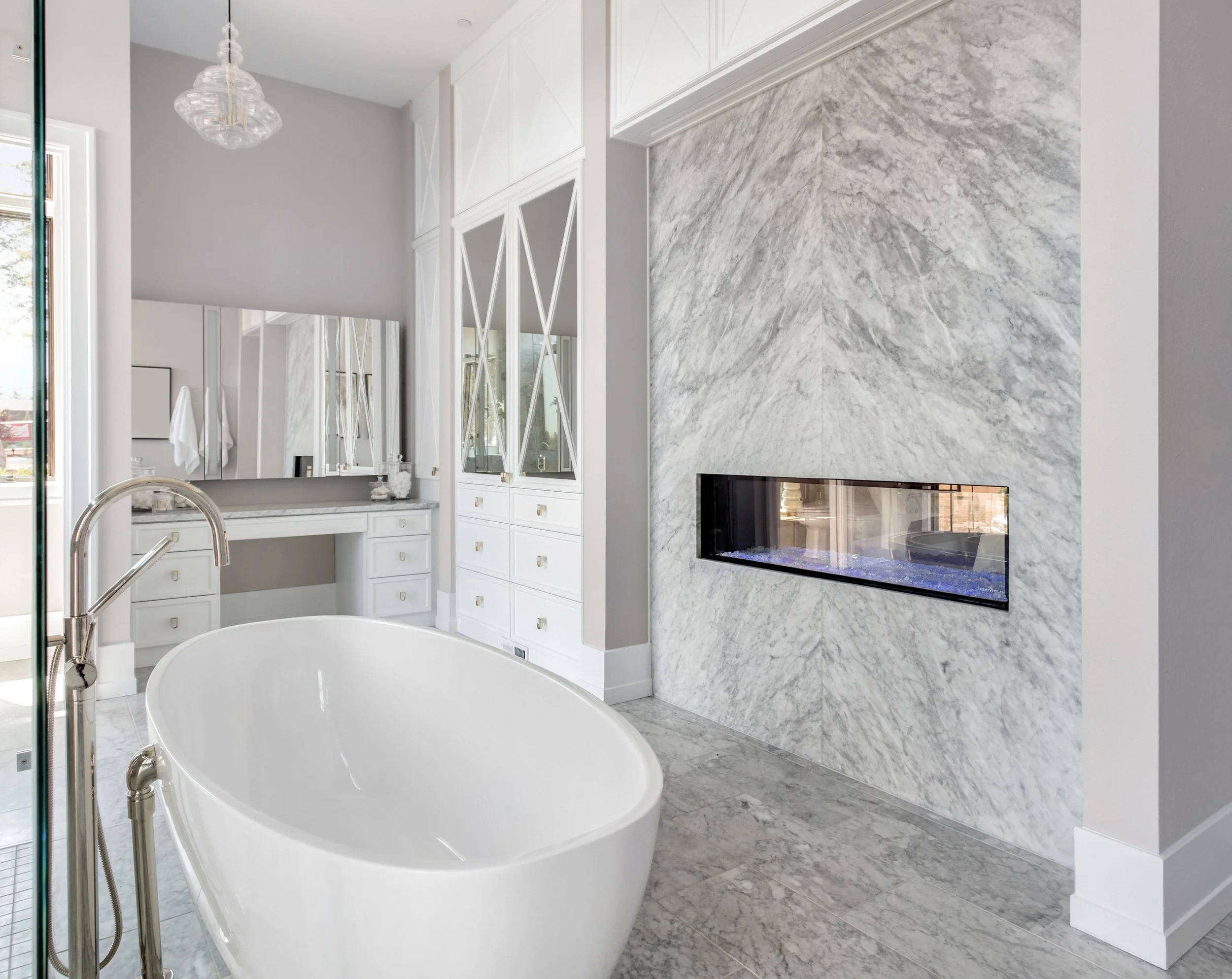 Modern bathroom with a white freestanding bathtub, marble flooring, and a marble accent wall with a built-in fireplace. There are white cabinets with gold handles and mirrors on the wall, with large windows providing natural light.
