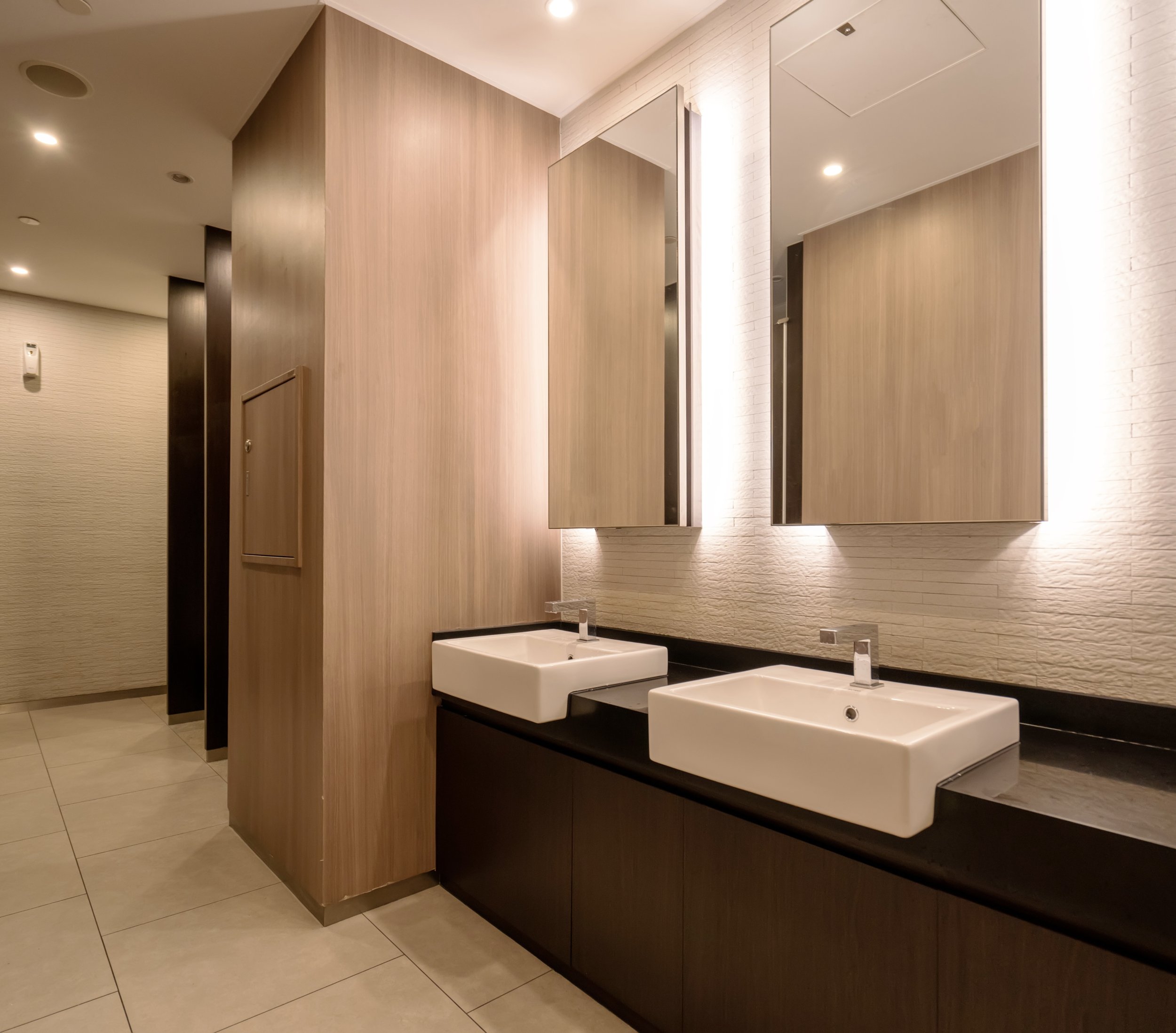 Public restroom with light wood and dark wood paneling, two white sinks with modern faucets, large mirror, beige tiled floor, white brick wall, ceiling with recessed lights.
