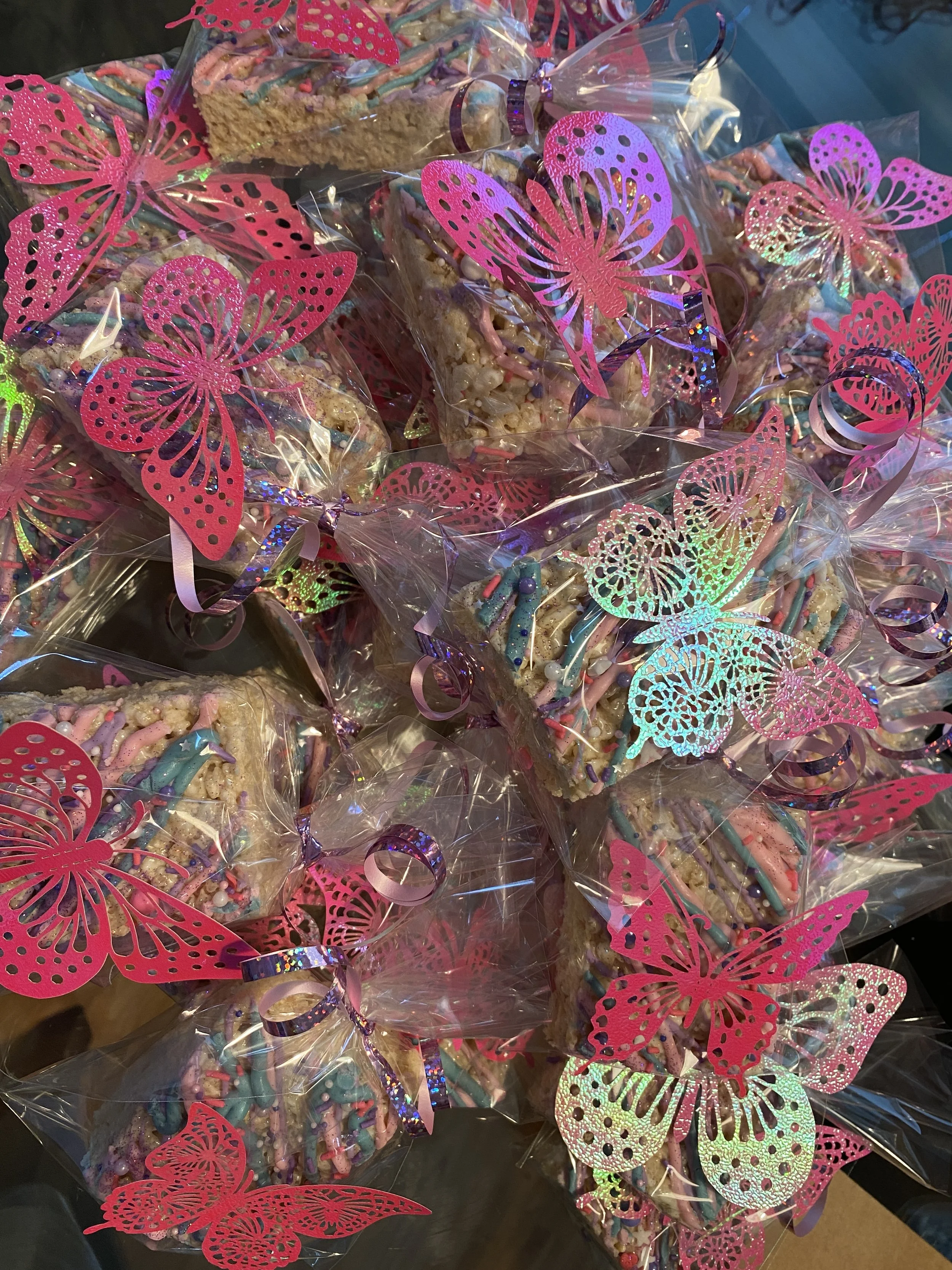 Multiple decorated rice crispy treats wrapped in clear plastic with pink butterfly-shaped paper cutouts and colorful sprinkles.