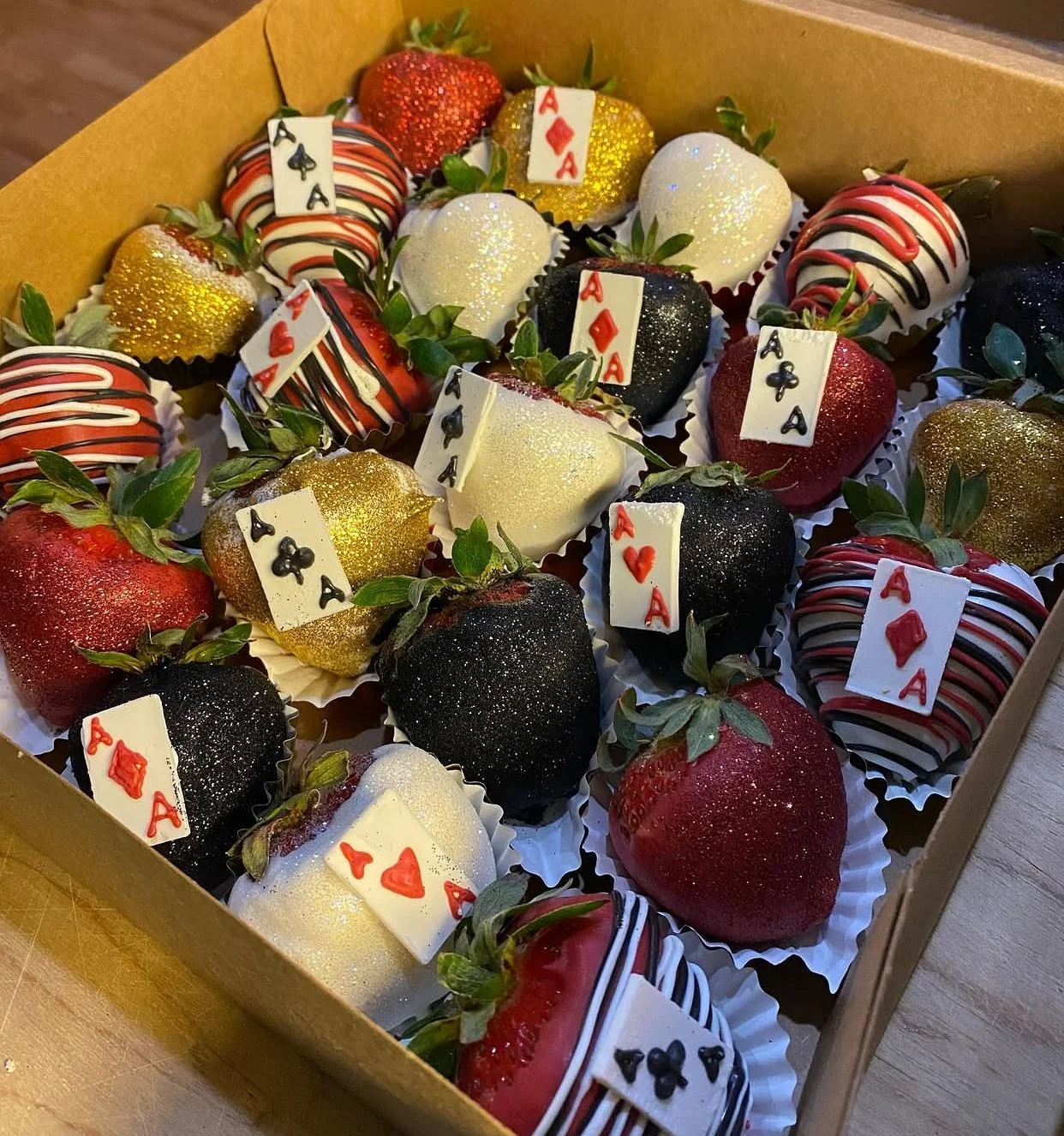 A box of decorated strawberries resembling playing cards, with some dipped in white, black, or red chocolate and topped with glitter or drizzle, and adorned with playing card symbols.