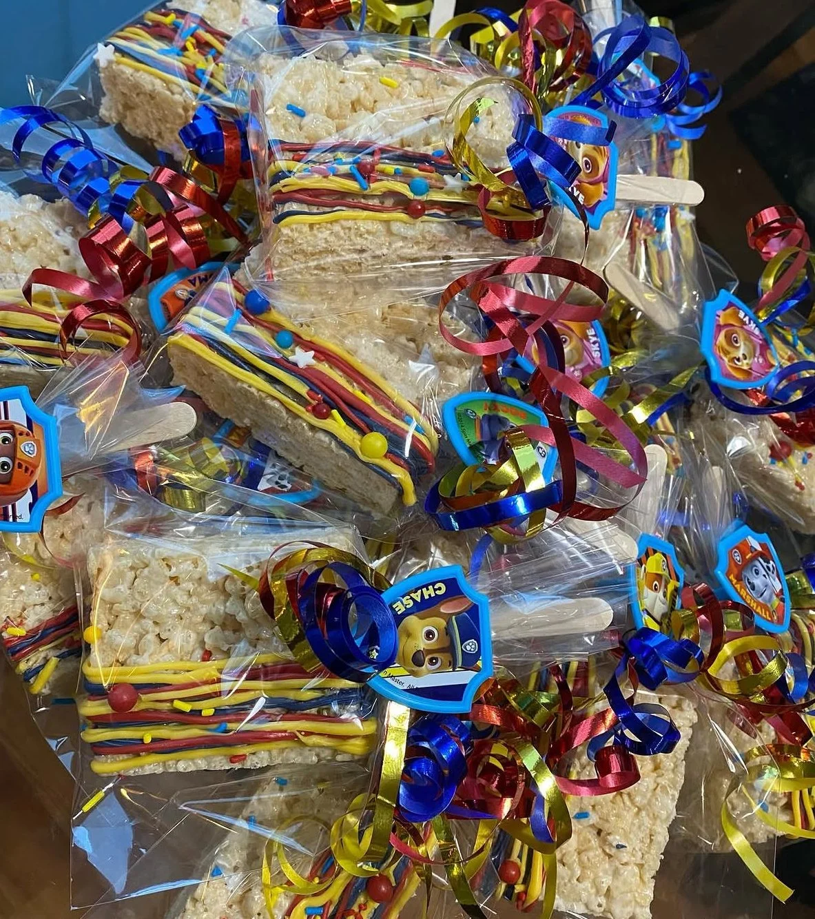 Chocolate rice crispy treats decorated with colorful icing and sprinkles, wrapped in clear plastic with confetti and a Paw Patrol badge sticker, intended for a celebration or party.