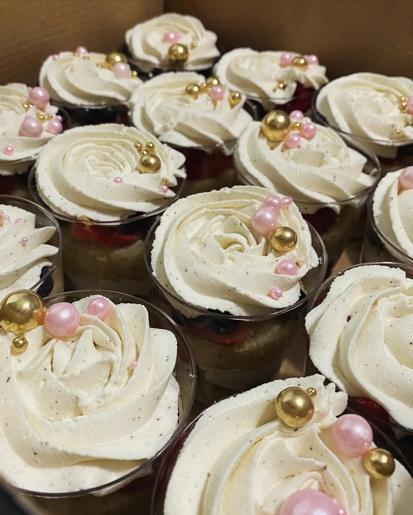 Multiple small dessert cups topped with swirls of white whipped cream or frosting, decorated with pink and gold spherical edible beads.