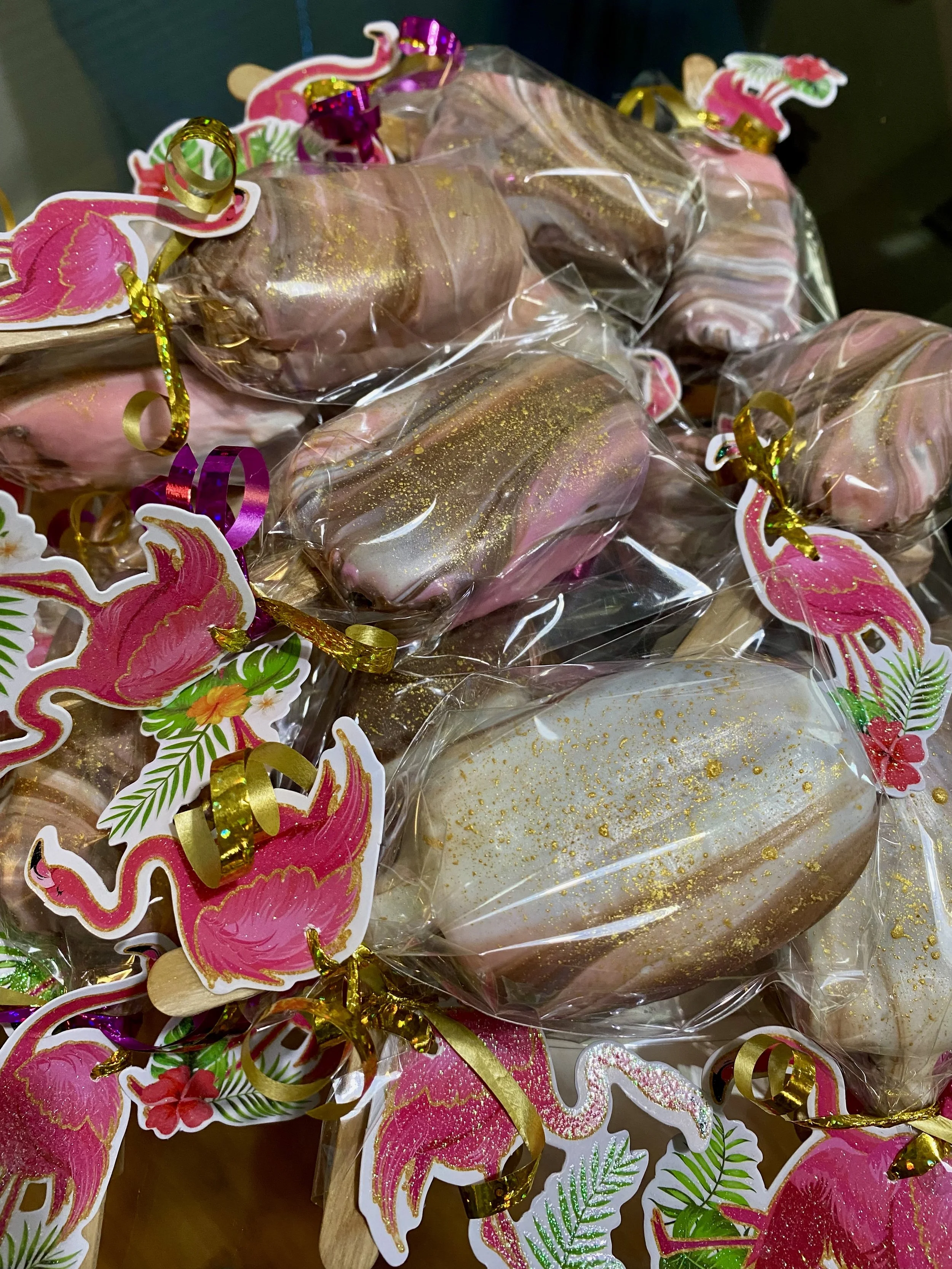 Multiple decorated marshmallow treats wrapped in clear plastic with pink flamingo and tropical leaf stickers, and gold and pink curl ribbons.