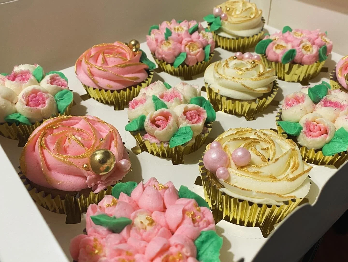 Assorted decorated cupcakes with pink, white, and cream frosting, topped with gold accents, small pearls, and green leaf decorations, arranged in a white box.