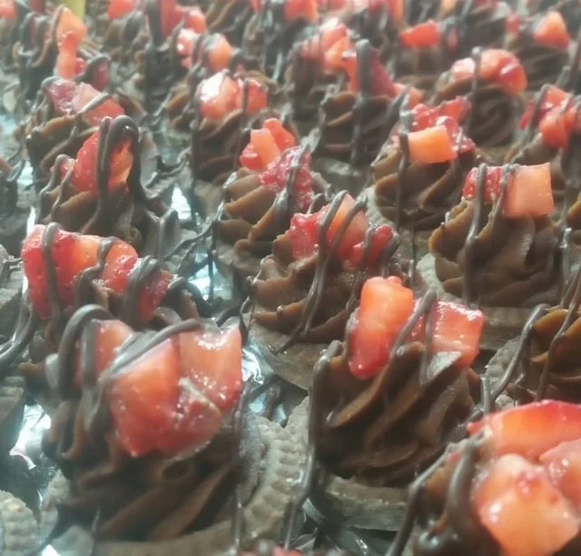 Multiple bite-sized chocolate desserts topped with diced strawberries and drizzled with chocolate, arranged on a metallic surface.