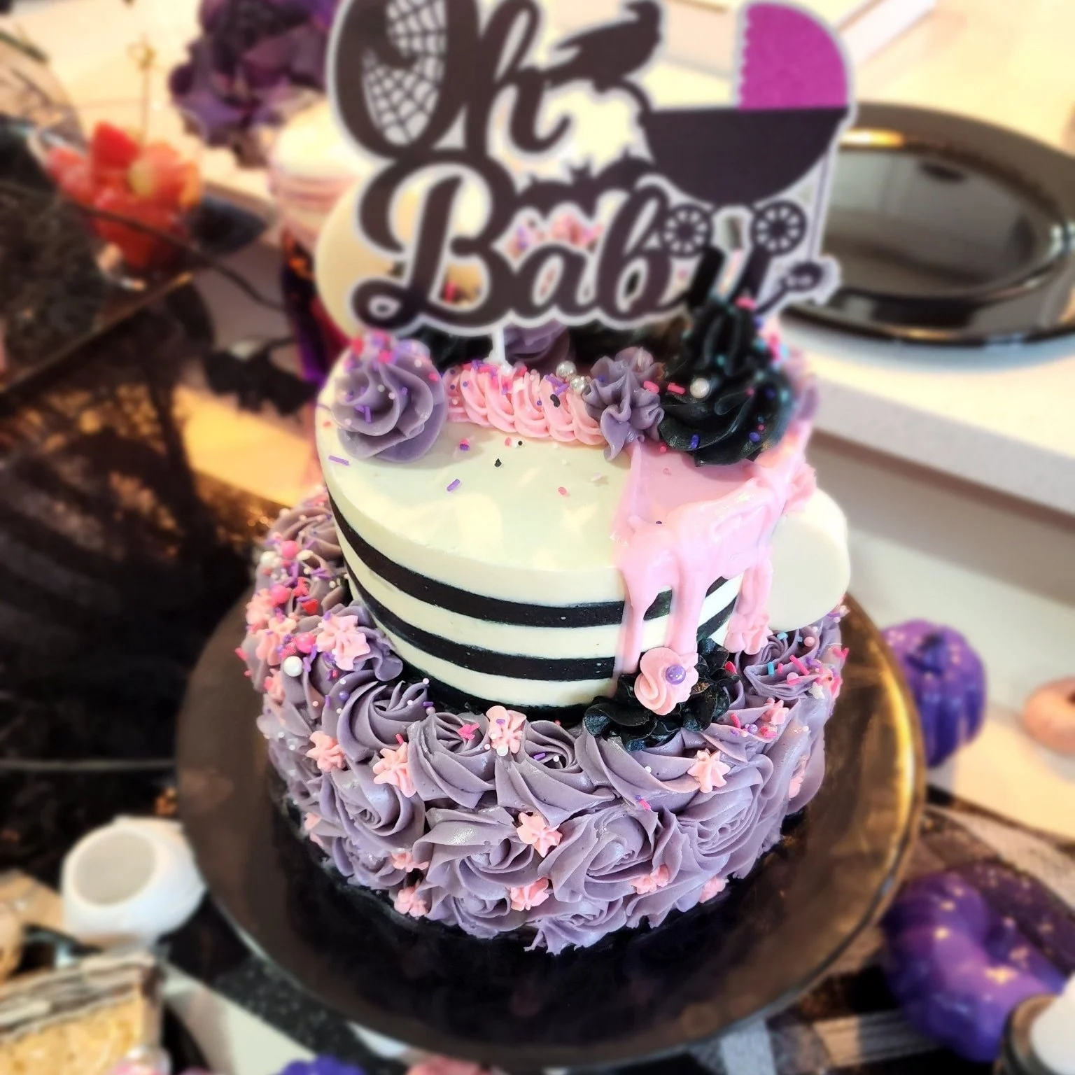 A purple and black striped birthday cake decorated with purple and pink cupcakes, pearl and sprinkles, topped with a sign that reads 'Happy Baby' with a pacifier motif, surrounded by purple and white decorations.
