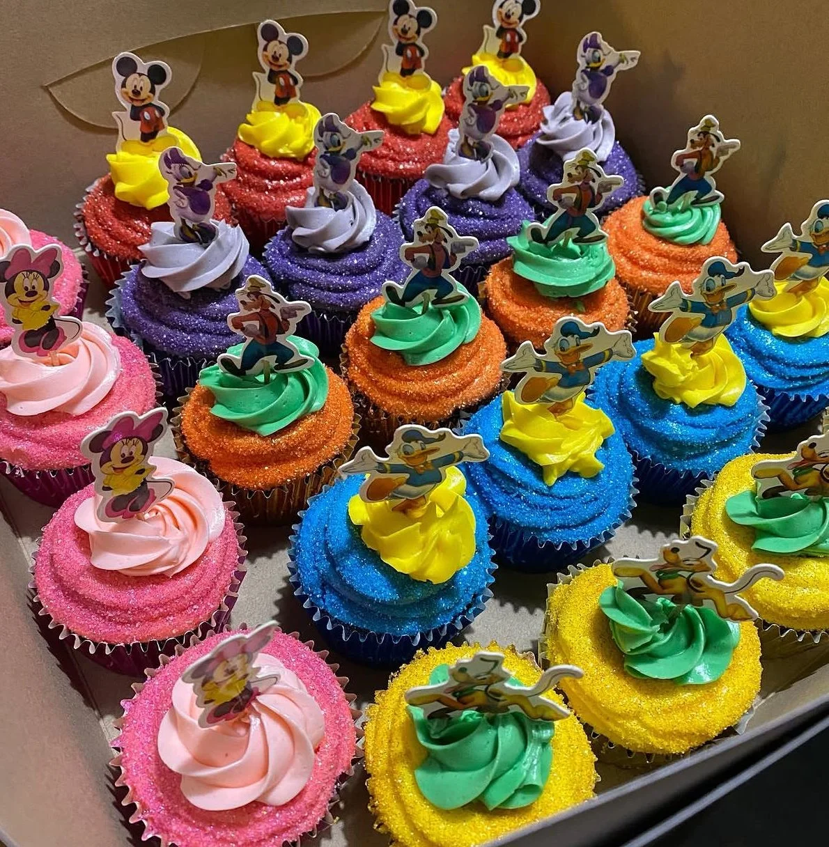 Colorful cupcakes decorated with Disney character toppers, including Mickey Mouse, Minnie Mouse, Donald Duck, and other Disney characters, with vibrant frosting in pink, purple, green, blue, orange, and yellow.