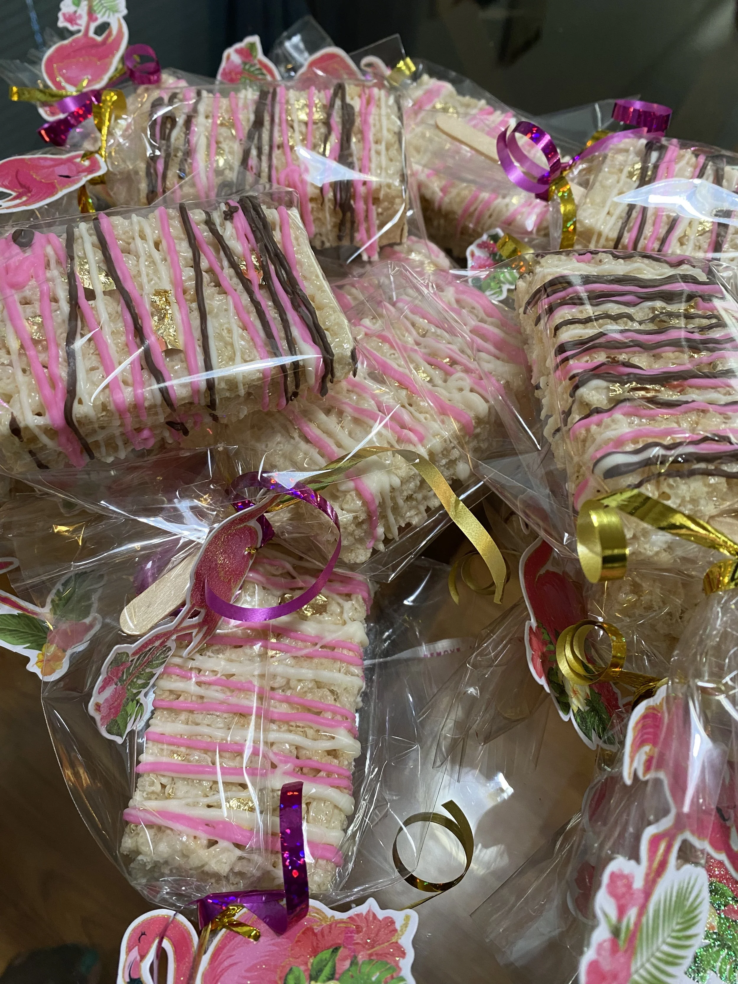 Rice Krispies treats decorated with pink, white, and chocolate drizzle, wrapped in clear plastic with colorful ribbons and flamingo-themed stickers.