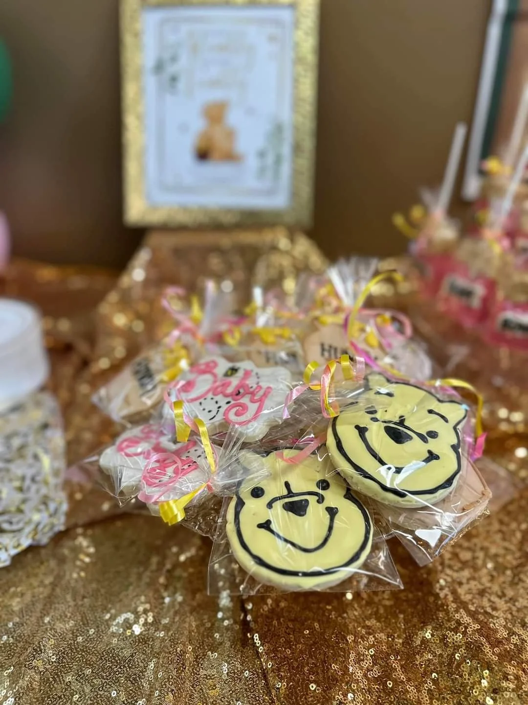 Close-up of decorated cookies featuring Winnie the Pooh faces, wrapped in clear plastic with pink and yellow ribbons, on a glittery gold tablecloth, with blurred background including a framed picture.
