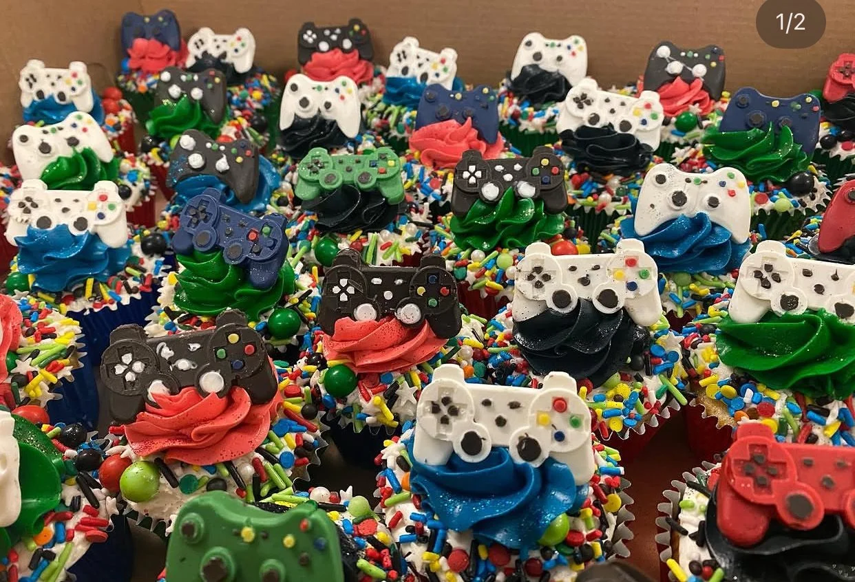 A tray of cupcakes decorated with colorful sprinkles, themed with gaming controllers and gaming-themed toppers.