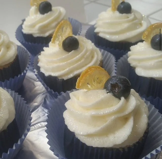 Cupcakes with white frosting, topped with a blueberry and a lemon slice, in blue paper liners.
