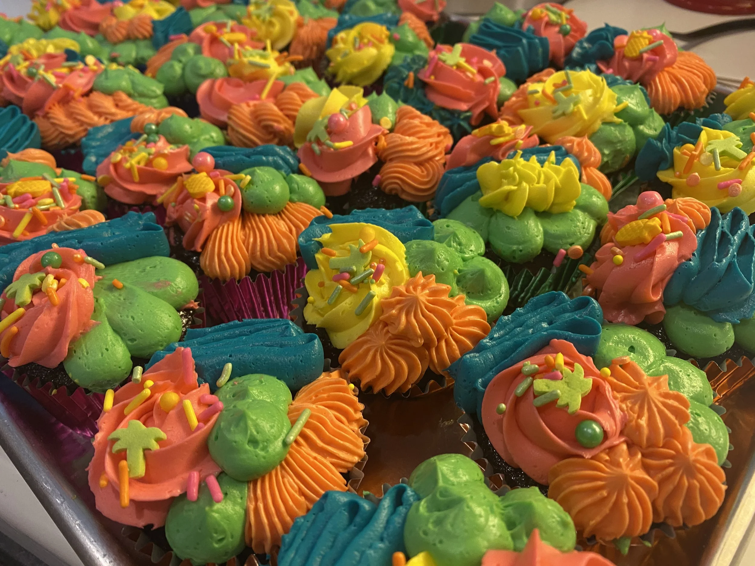 Colorful cupcakes decorated with bright pink, yellow, green, and blue icing topped with sprinkles and small edible decorations, arranged on a silver tray.