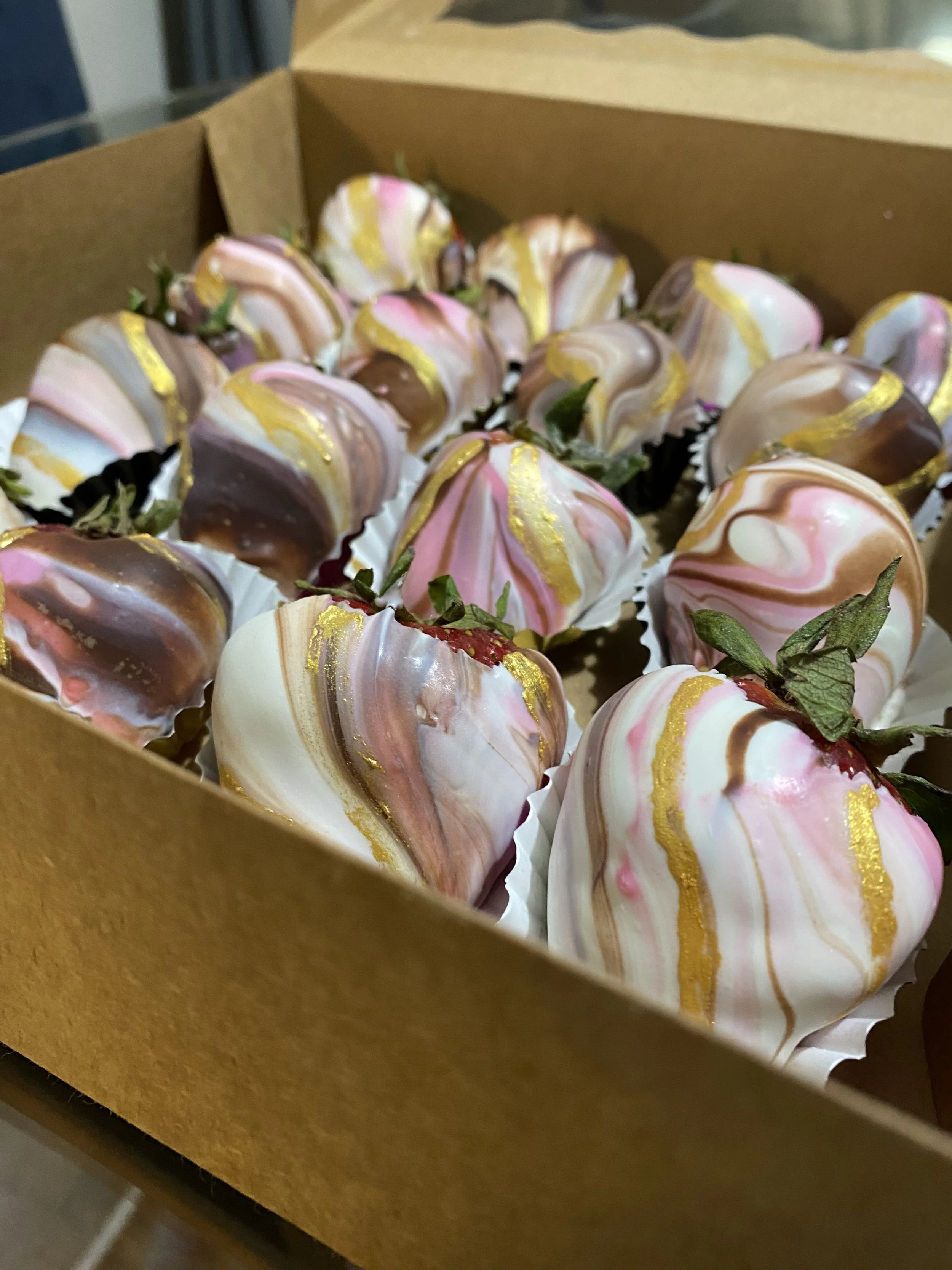Close-up of strawberries dipped in white, pink, and brown chocolate with gold accents, arranged in a cardboard box.