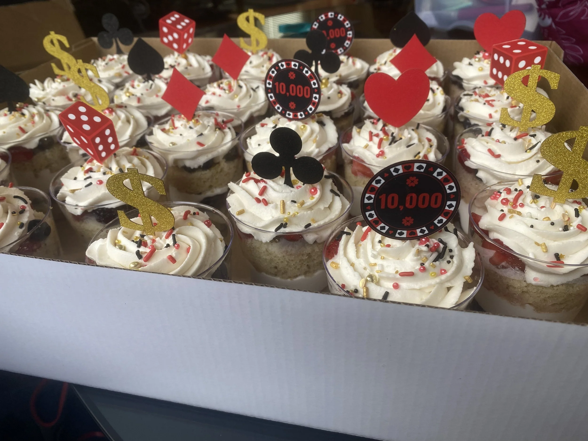 A box of cupcakes decorated with white frosting, sprinkles, and casino-themed toppers such as gold dollar signs, red hearts, black spades, and red diamonds, along with red dice and black poker chips.