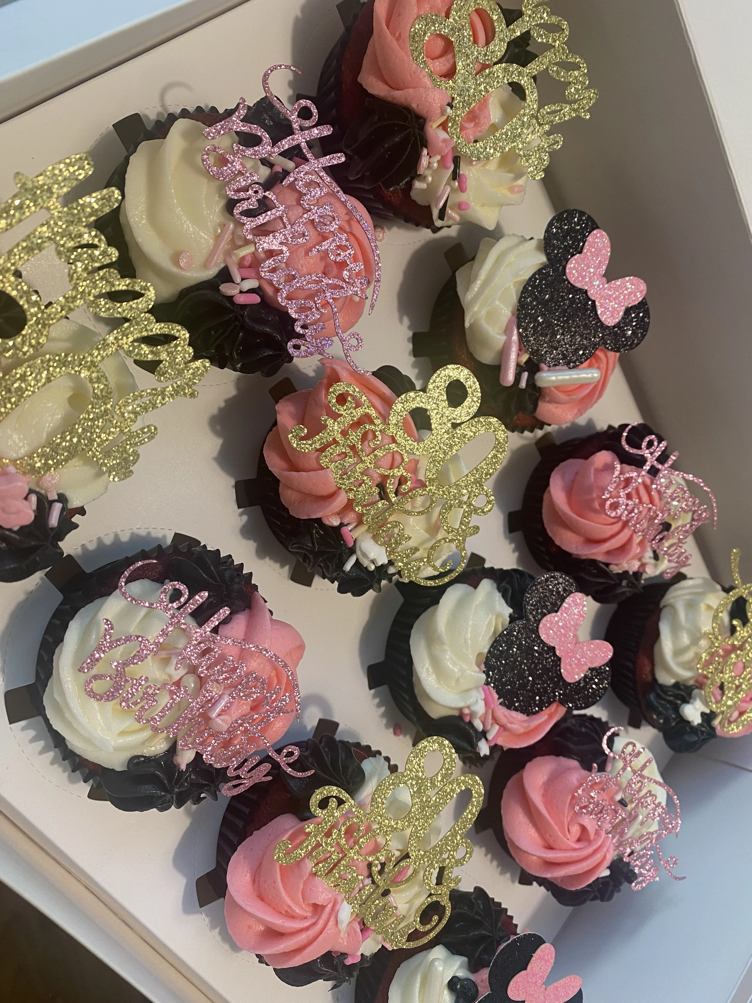 Assorted cupcakes decorated with pink, white, black, and glittery edible toppers, including hearts, bows, and 'happy birthday' signs, arranged in a white box.