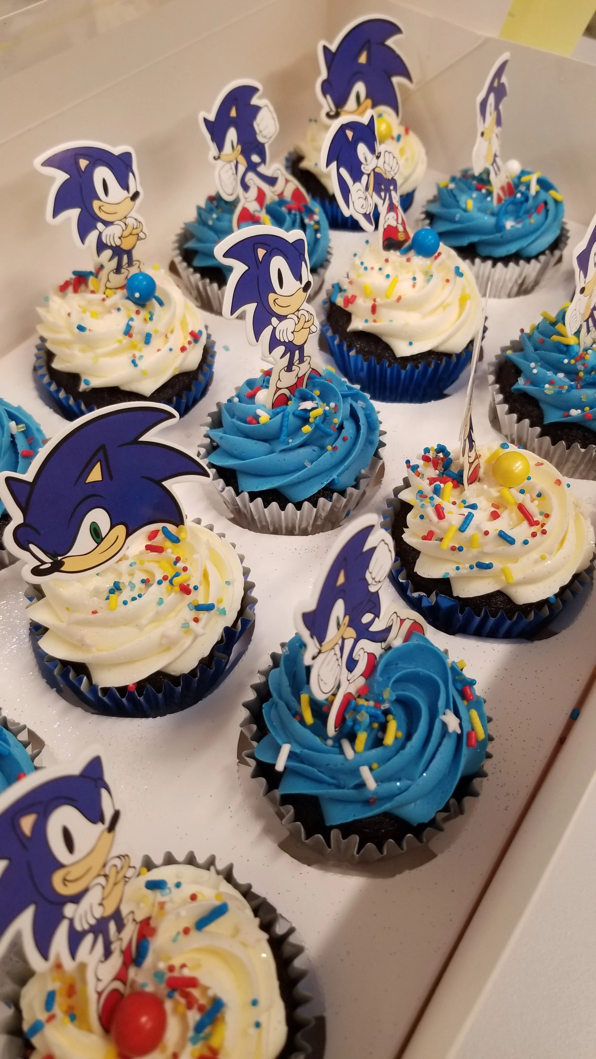 Decorated cupcakes featuring Sonic the Hedgehog toppers, with blue and white icing, rainbow sprinkles, and a cherry on some.