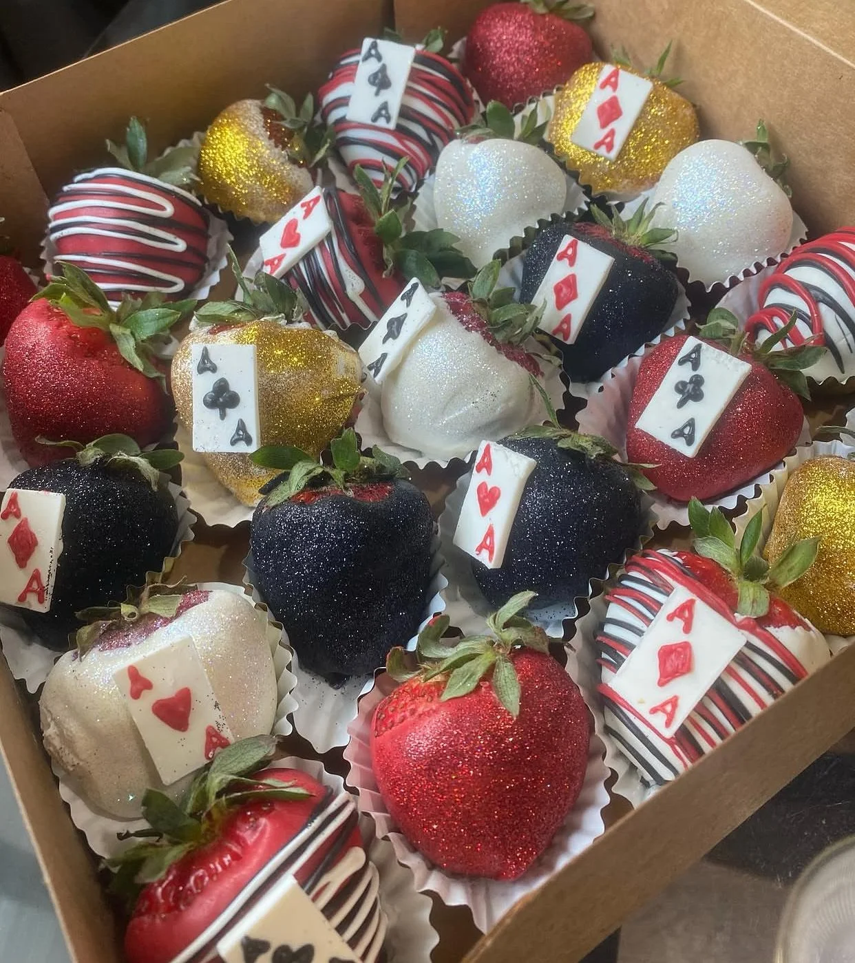 A box of strawberries decorated to look like playing cards, featuring red, black, white, gold, and glitter. Some strawberries are plain, while others have drizzles or edible decorations resembling poker hands like aces and queens.