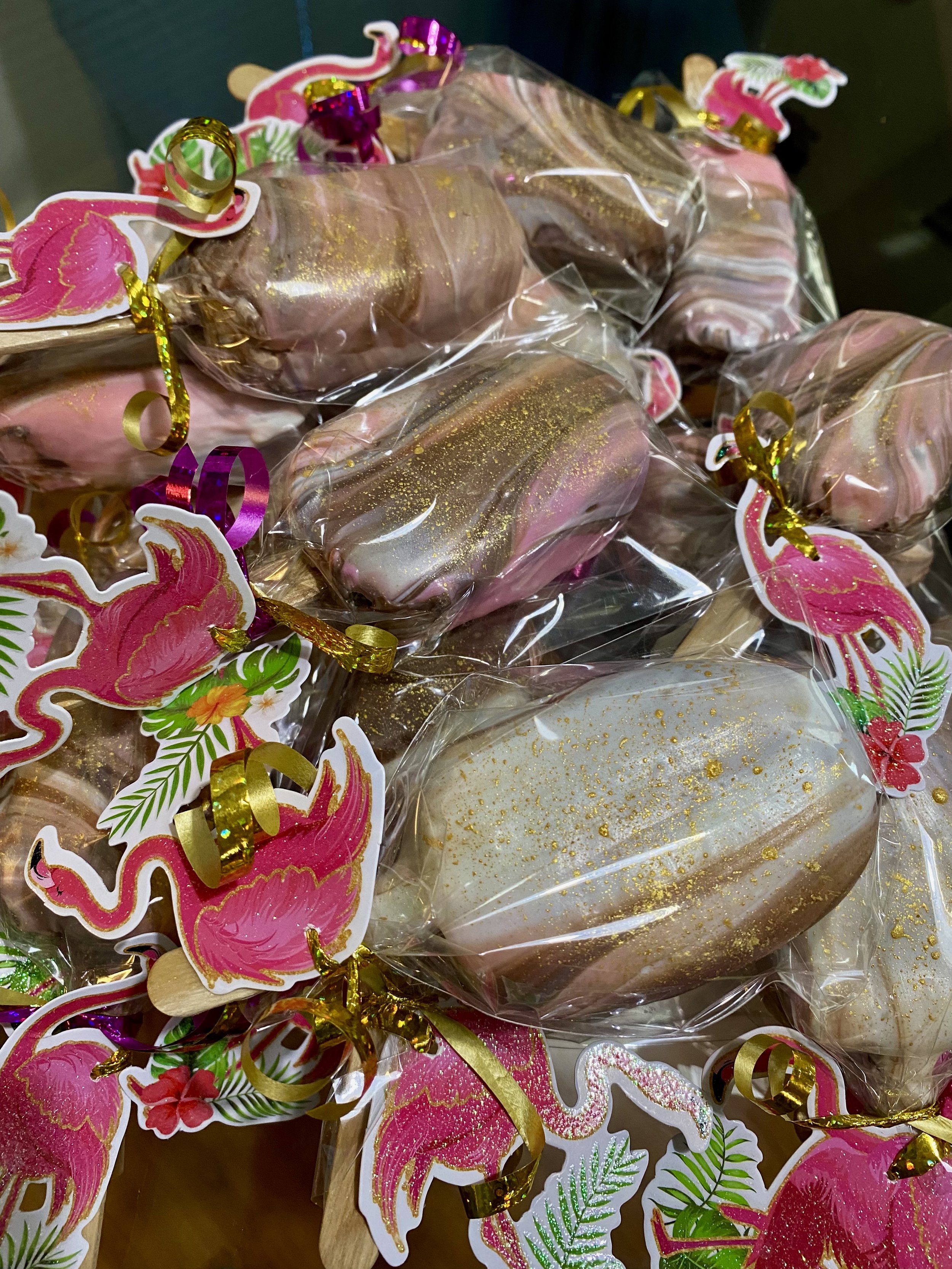 Decorative pink flamingo stickers with green palm leaves and gold glitter, attached to clear plastic wrapping around baked goods or confections.