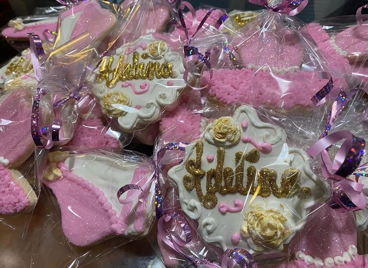 Various decorated cookies wrapped in clear plastic with pink wrapping ribbons, including cookies with 'Mama' and 'Daughter' written in gold, and pink and white themed designs, likely for a celebration.