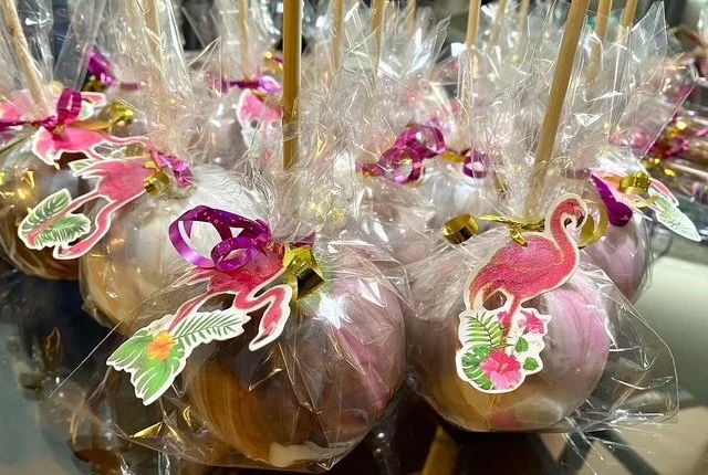 Chocolate apples decorated with flamingo and tropical leaf stickers, wrapped in clear plastic and tied with colorful ribbons, on skewers.