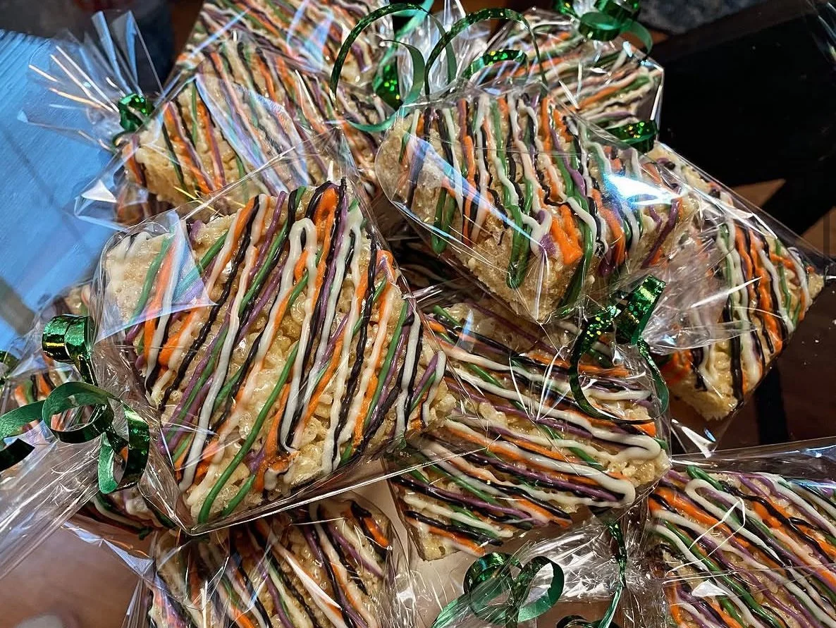 A pile of packaged rice krispie treats decorated with colorful frosting and drizzled with icing, wrapped in clear plastic and tied with green ribbons.