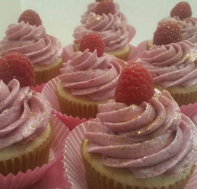 A group of cupcakes with pink frosting, topped with raspberries and gold glitter, in pink cupcake liners.