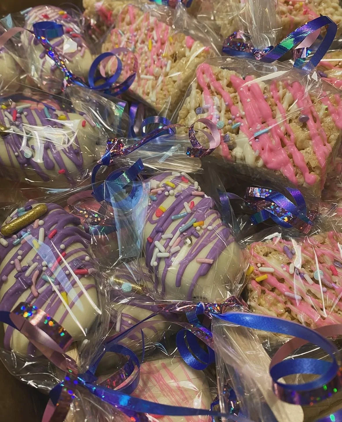 Assorted decorated cakes or cookies wrapped in clear plastic with pink, white, and purple icing, sprinkles, and topped with blue and pink ribbons.