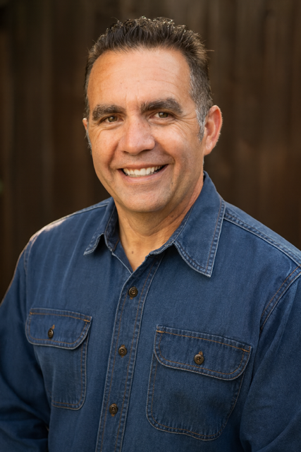 A smiling middle-aged man with short dark hair, wearing a denim shirt, standing in front of a wooden background.