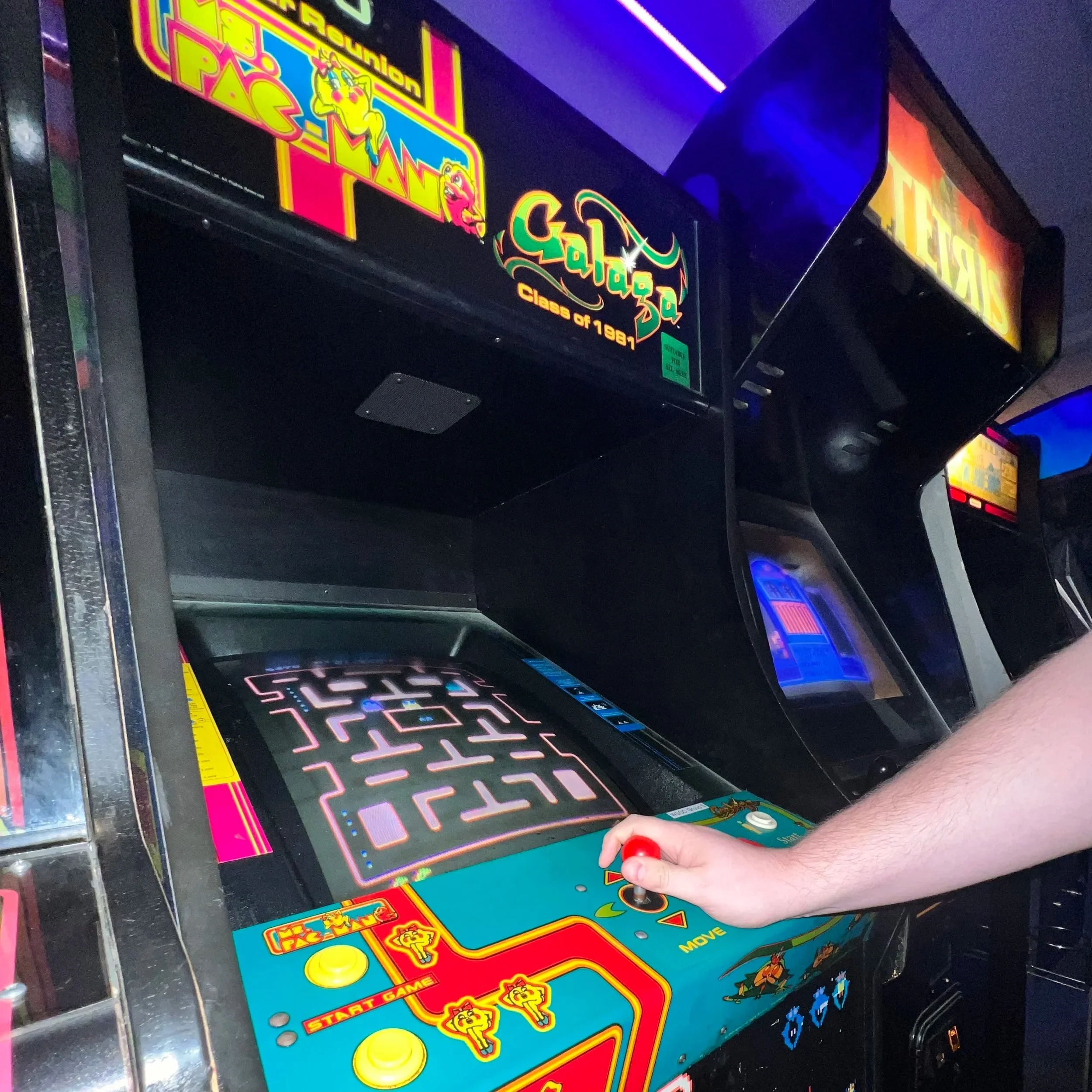 A person playing Pac-Man on an arcade machine in a dimly lit arcade. The cabinet features colorful graphics and neon-themed decorations.