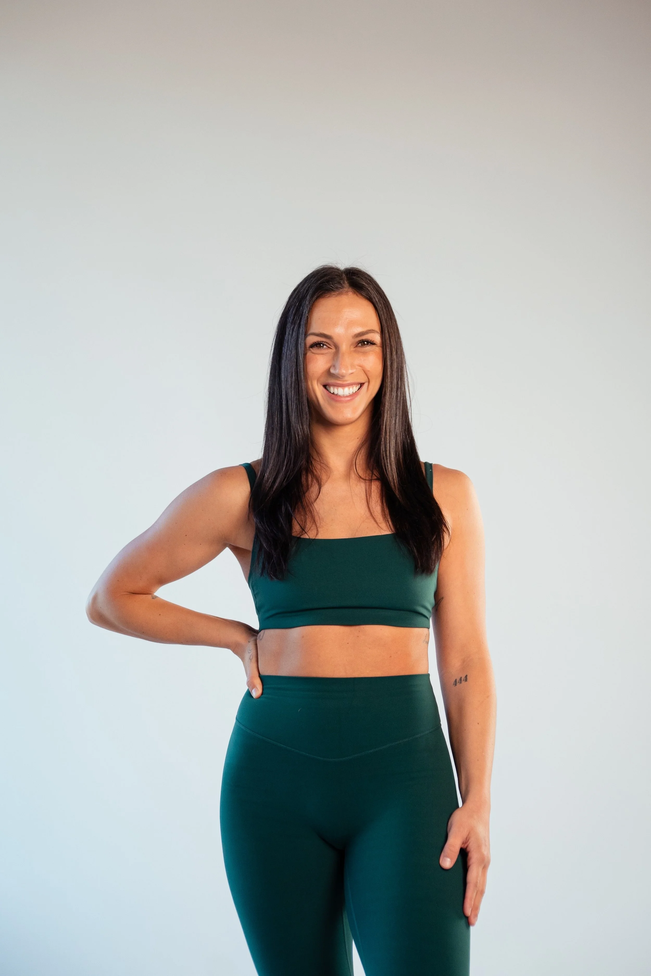A woman with long dark hair, wearing a dark green sports bra and matching high-waisted leggings, standing against a plain light background.