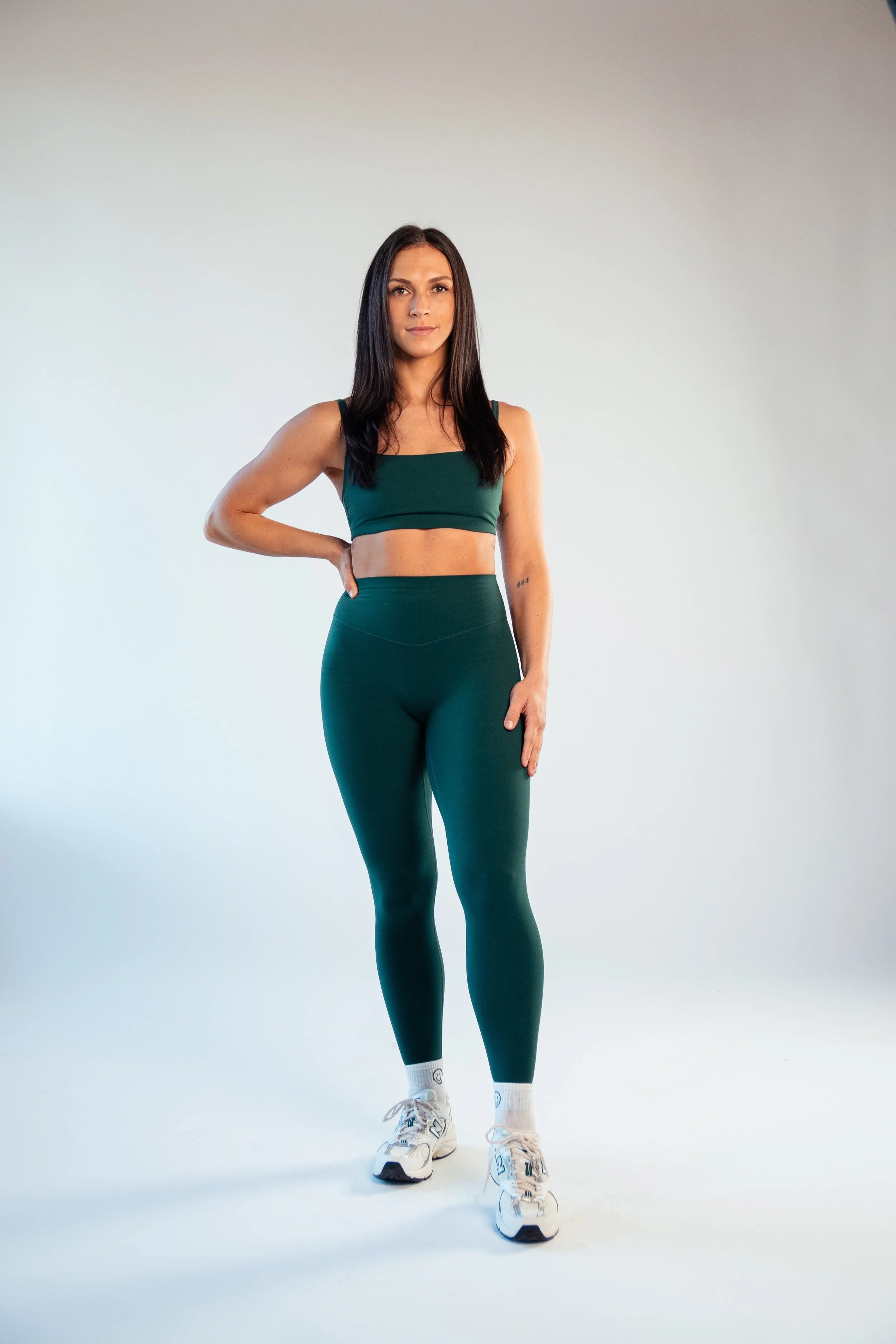 A woman with long dark hair, wearing a dark green sports bra and matching high-waisted leggings, standing against a plain light background.