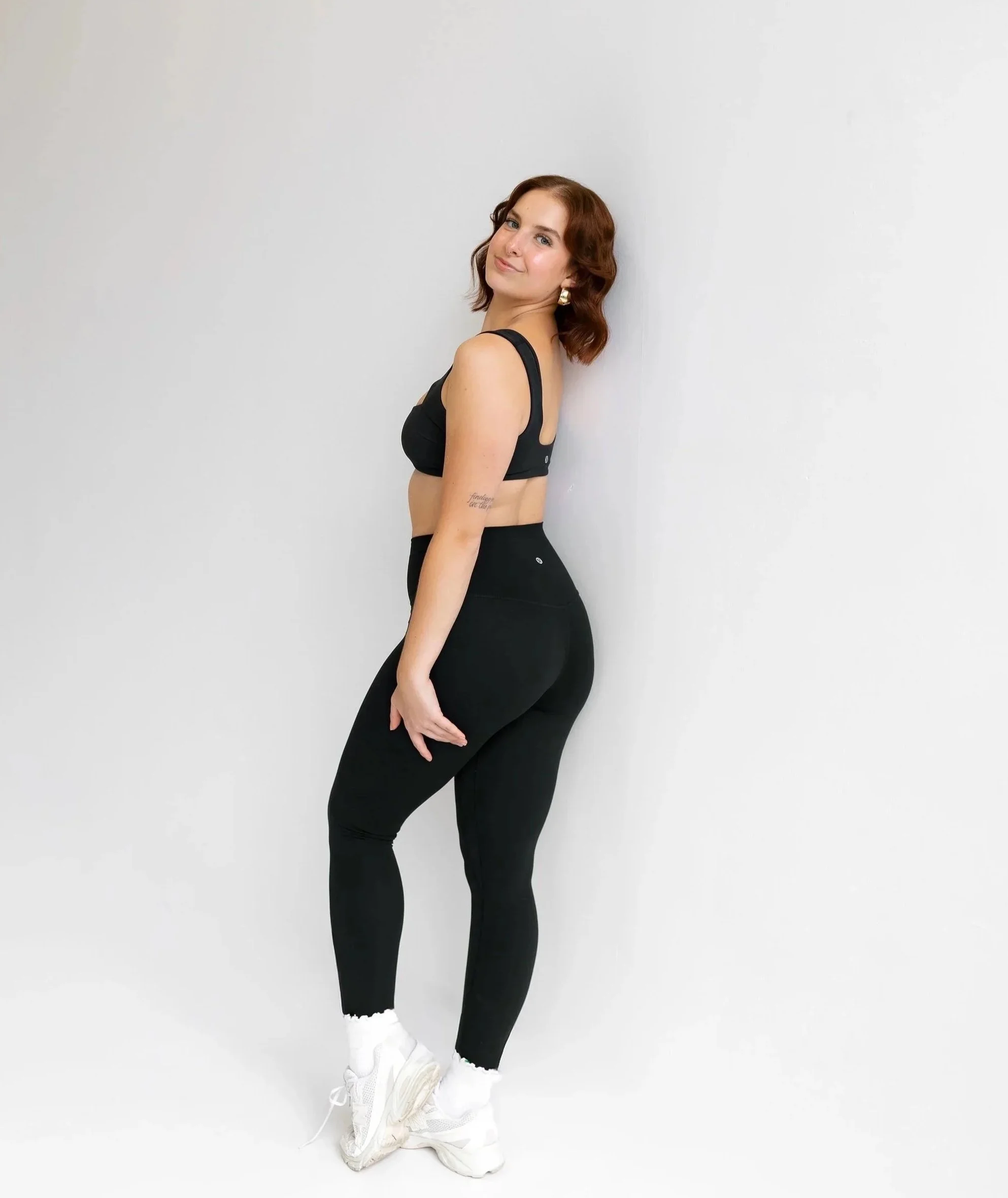 A woman in red athletic wear and gold earrings, with a gray sweatshirt slung over her shoulders, standing against a plain white wall.