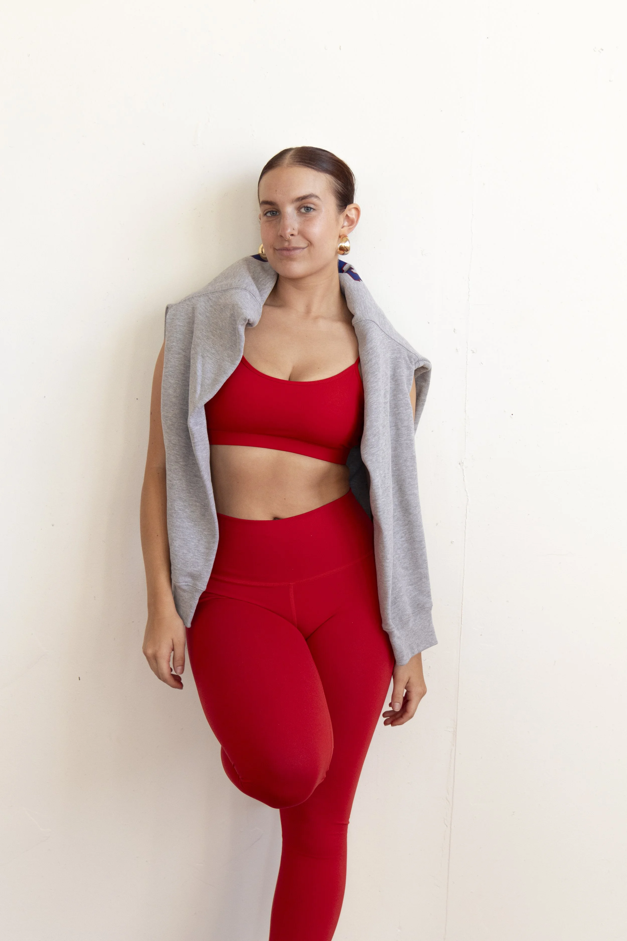 A woman in red athletic wear and gold earrings, with a gray sweatshirt slung over her shoulders, standing against a plain white wall.