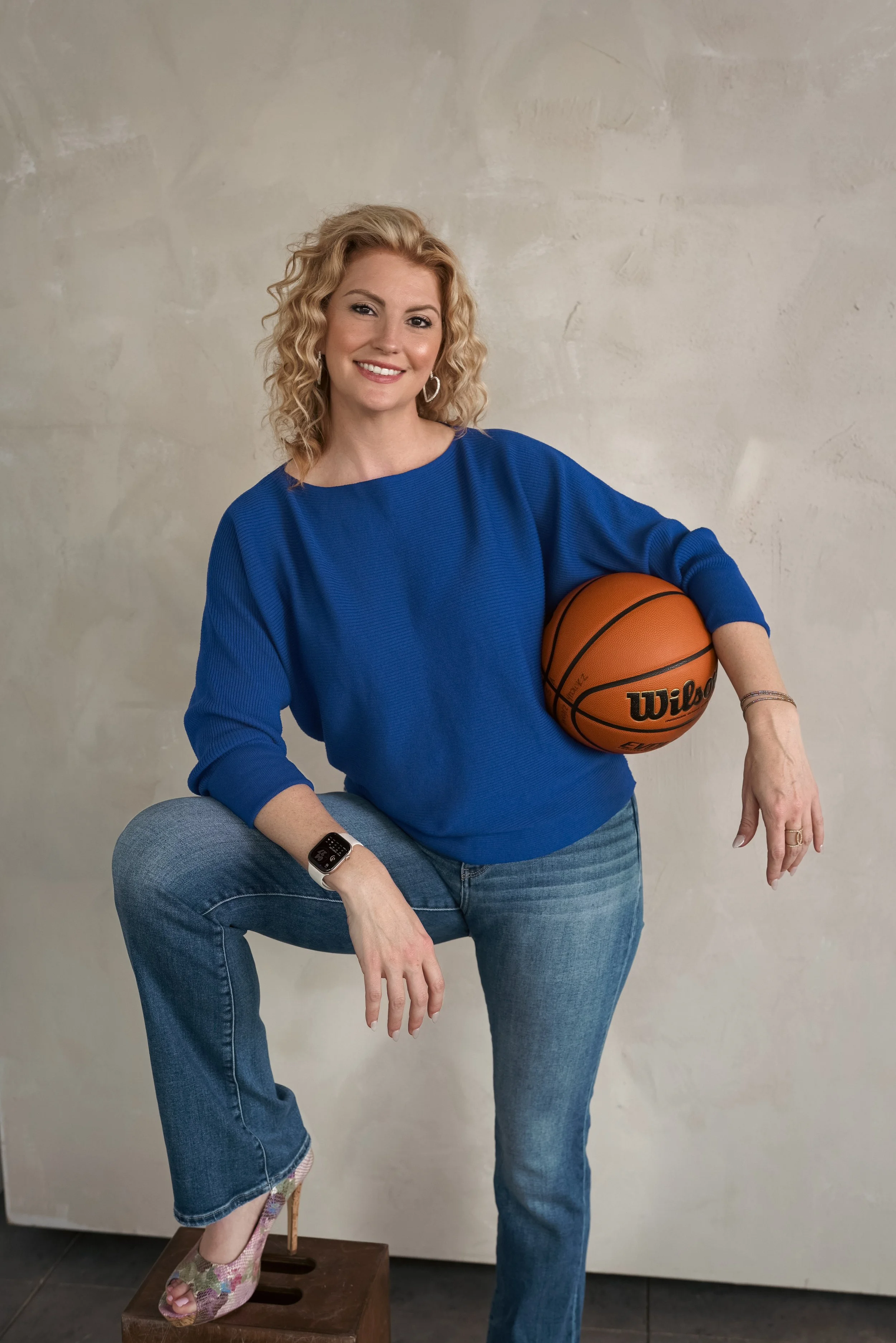 A woman with blonde curly hair wearing a blue top, jeans, and colorful high heels, holding an orange Wilson basketball, and smiling, positioned with one foot on a wooden step stool against a beige wall.