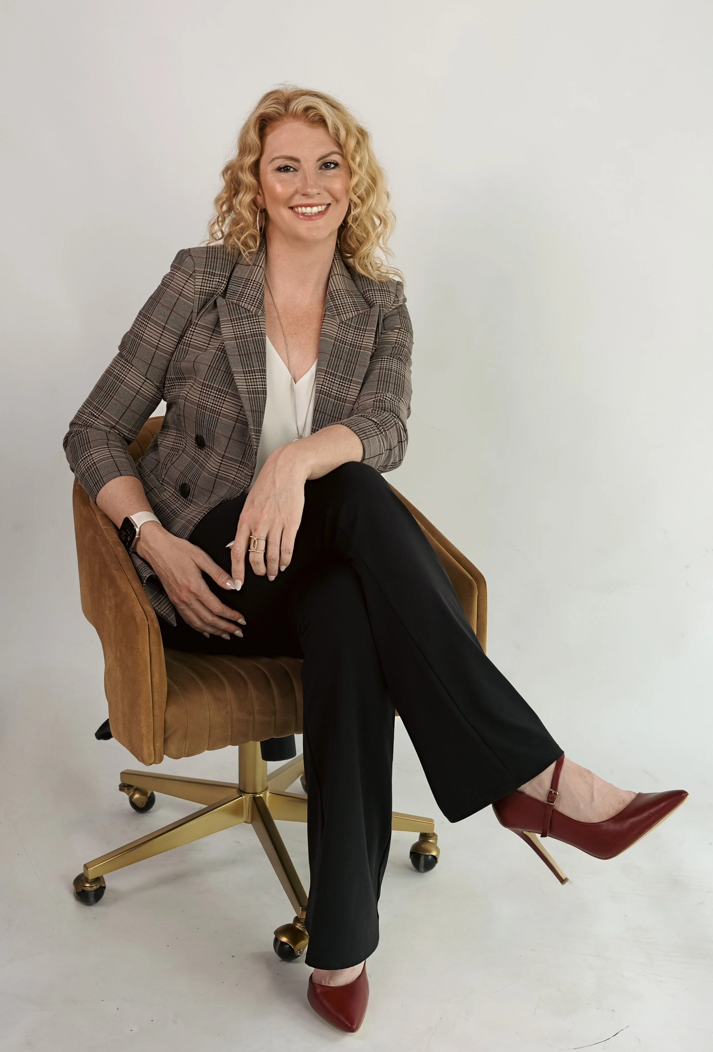 A woman with blonde curly hair wearing a plaid blazer, white blouse, black pants, and red high heels sitting on a brown chair with wheels against a plain white background.