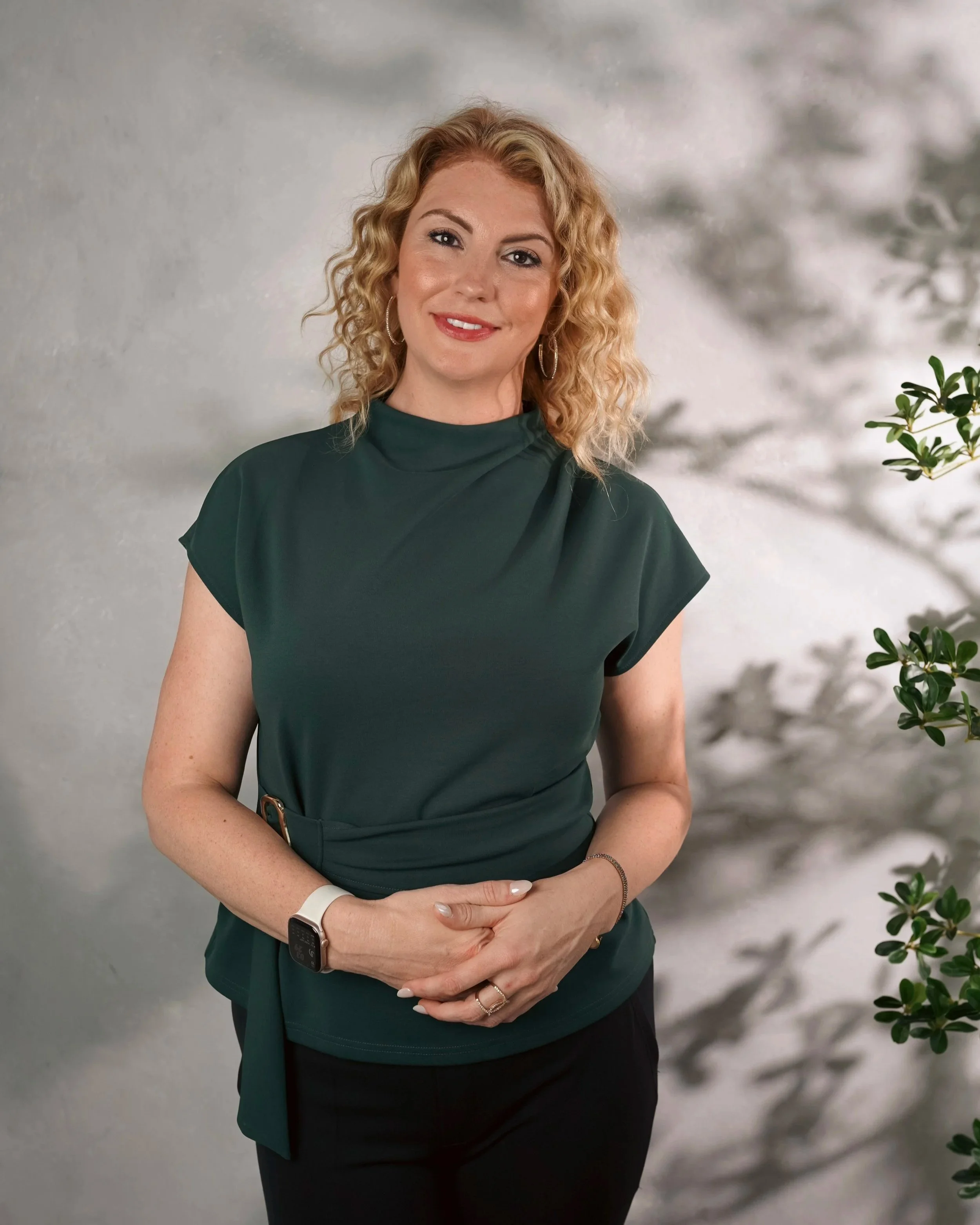 A woman with blonde curly hair and hoop earrings standing in front of a blurred gray background with some greenery on the side. She is wearing a green dress and smiling at the camera.