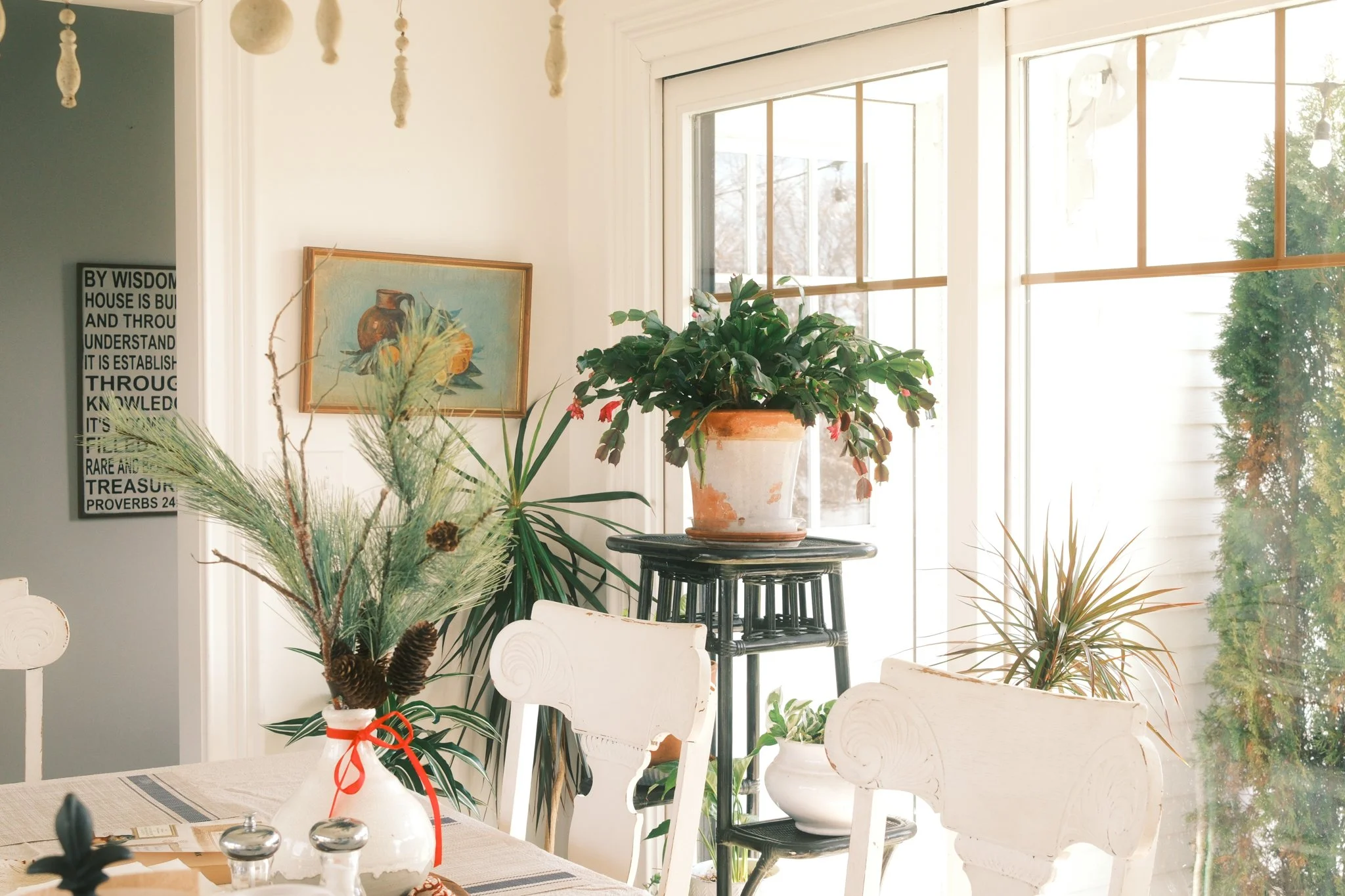 Bright dining or living area with large window, potted plants, artwork, and decorative objects on tables and shelves.
