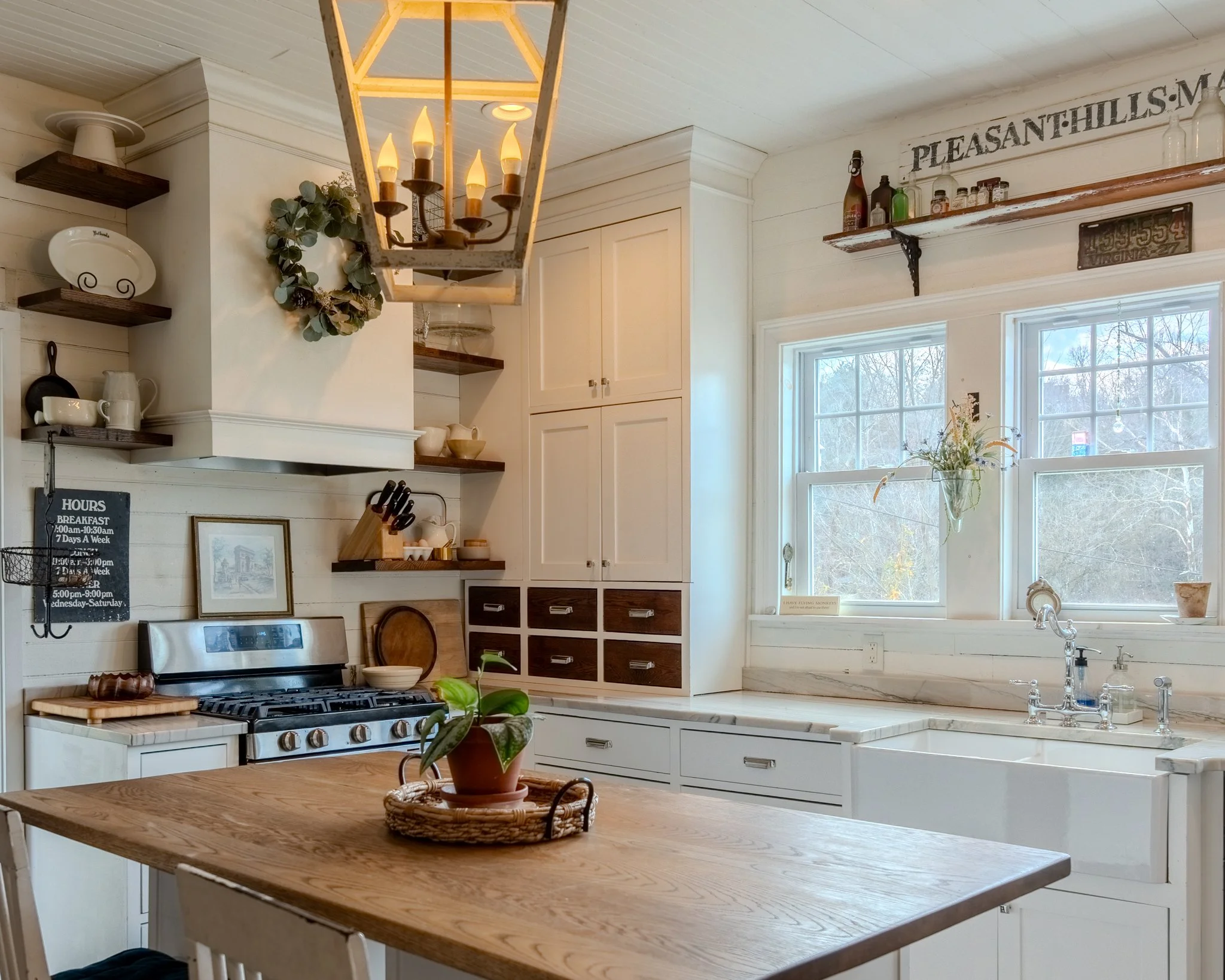 A cozy kitchen with white cabinets, a wooden island, open wooden shelves with decorative dishes, a stove with a plant in a pot on the counter, and a large window with a view of trees outside. A hanging light fixture with candle-shaped bulbs is above the island.