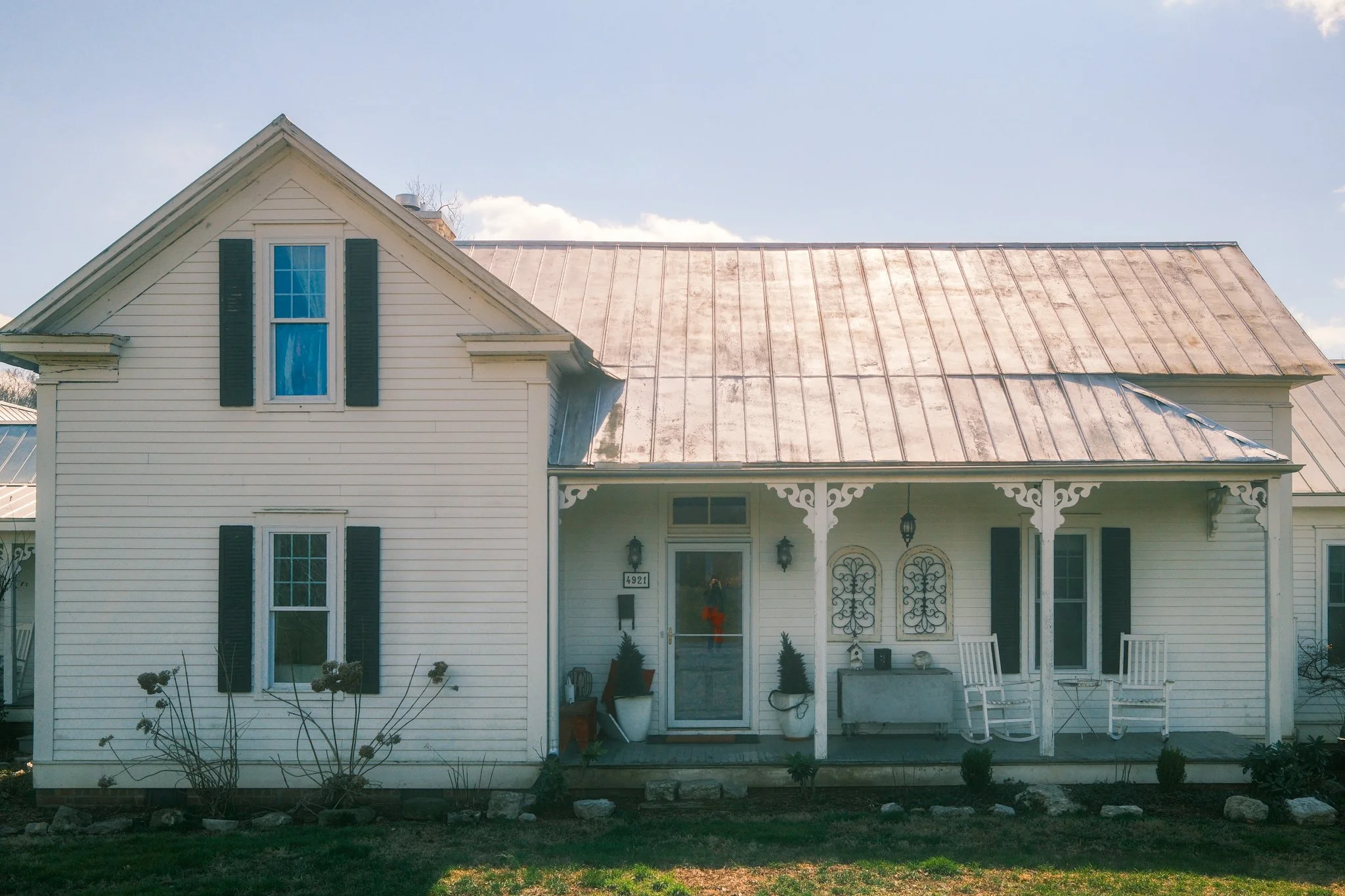 Front view of a white house with black shutters and a metal roof, decorated with fall decorations, in a suburban neighborhood.
