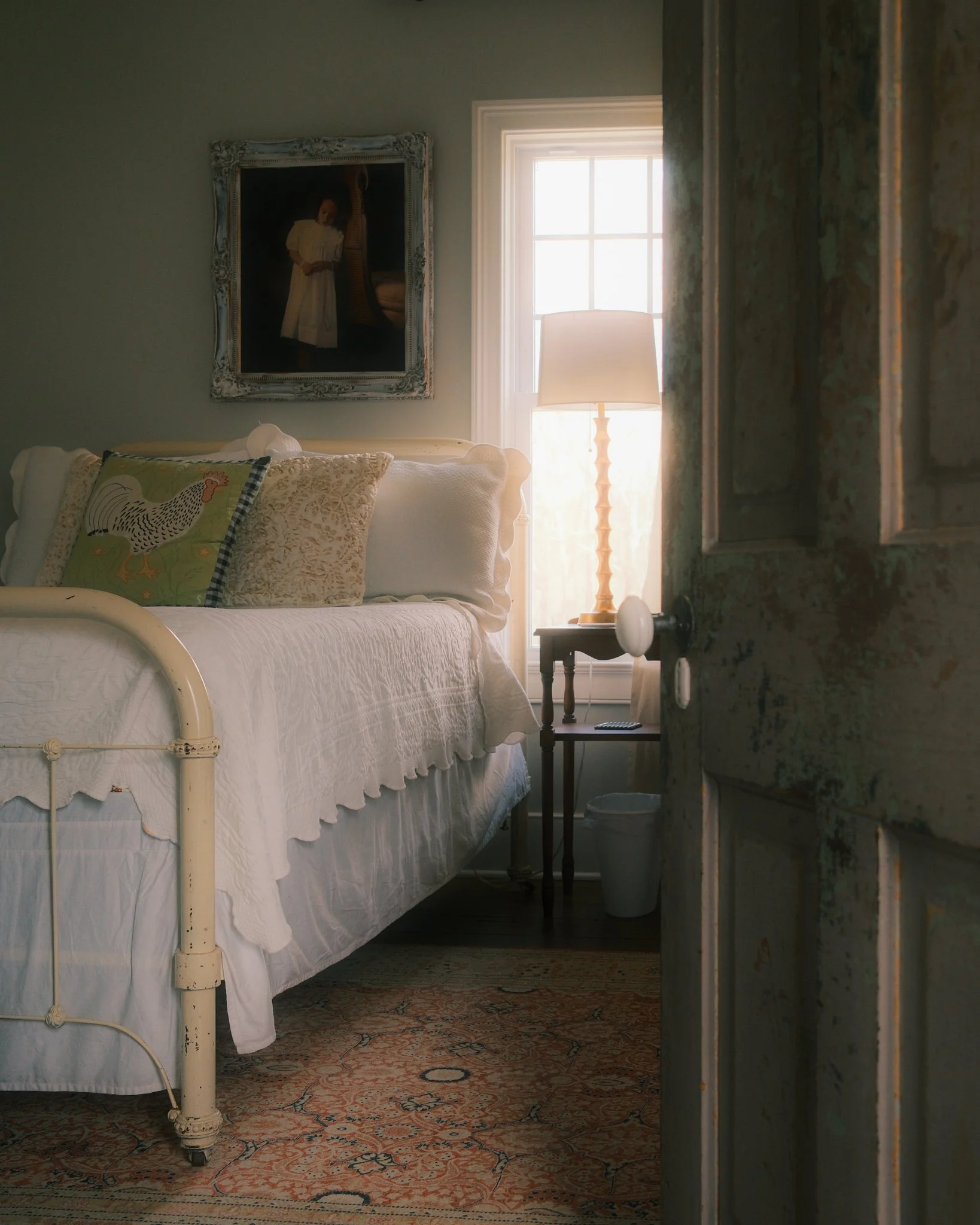 A bedroom seen through a partially opened distressed wooden door with a visible hinges. Inside, the bed has a white quilt and pillows, including one with a hen design. There is a side table holding a lamp with a cream-colored shade and a small object. A framed portrait of a woman in a white dress hangs on the wall, with a window allowing natural light to illuminate the room.