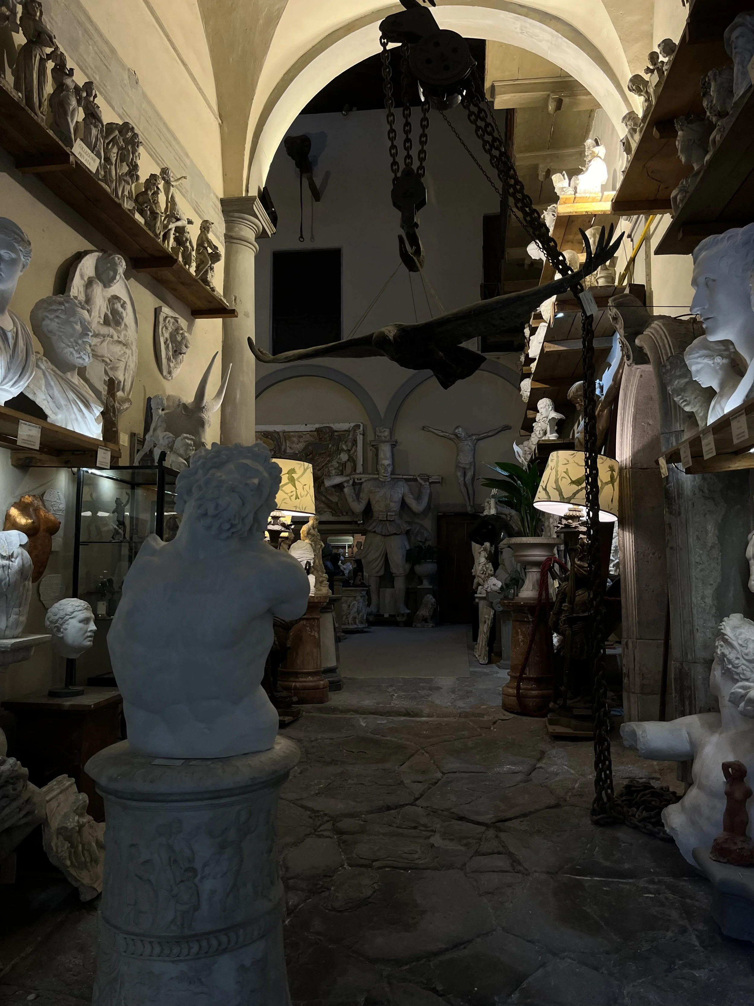 An art studio filled with classical bust sculptures, statues, and wall carvings, with a large hanging bird sculpture and dark stone floor.