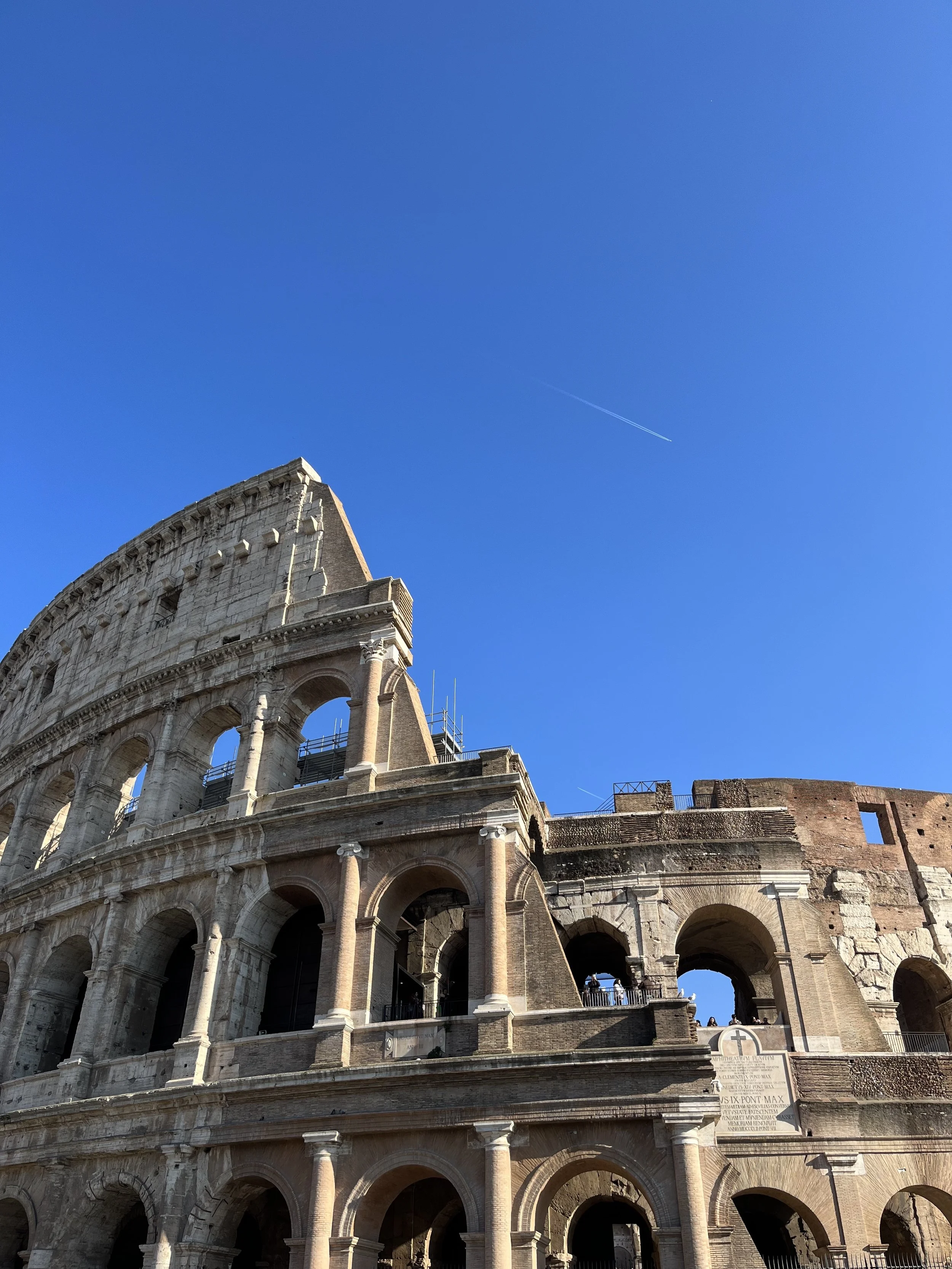 Close-up of the exterior of the Colosseum in Rome, Italy, against a bright blue sky with a faint contrail.