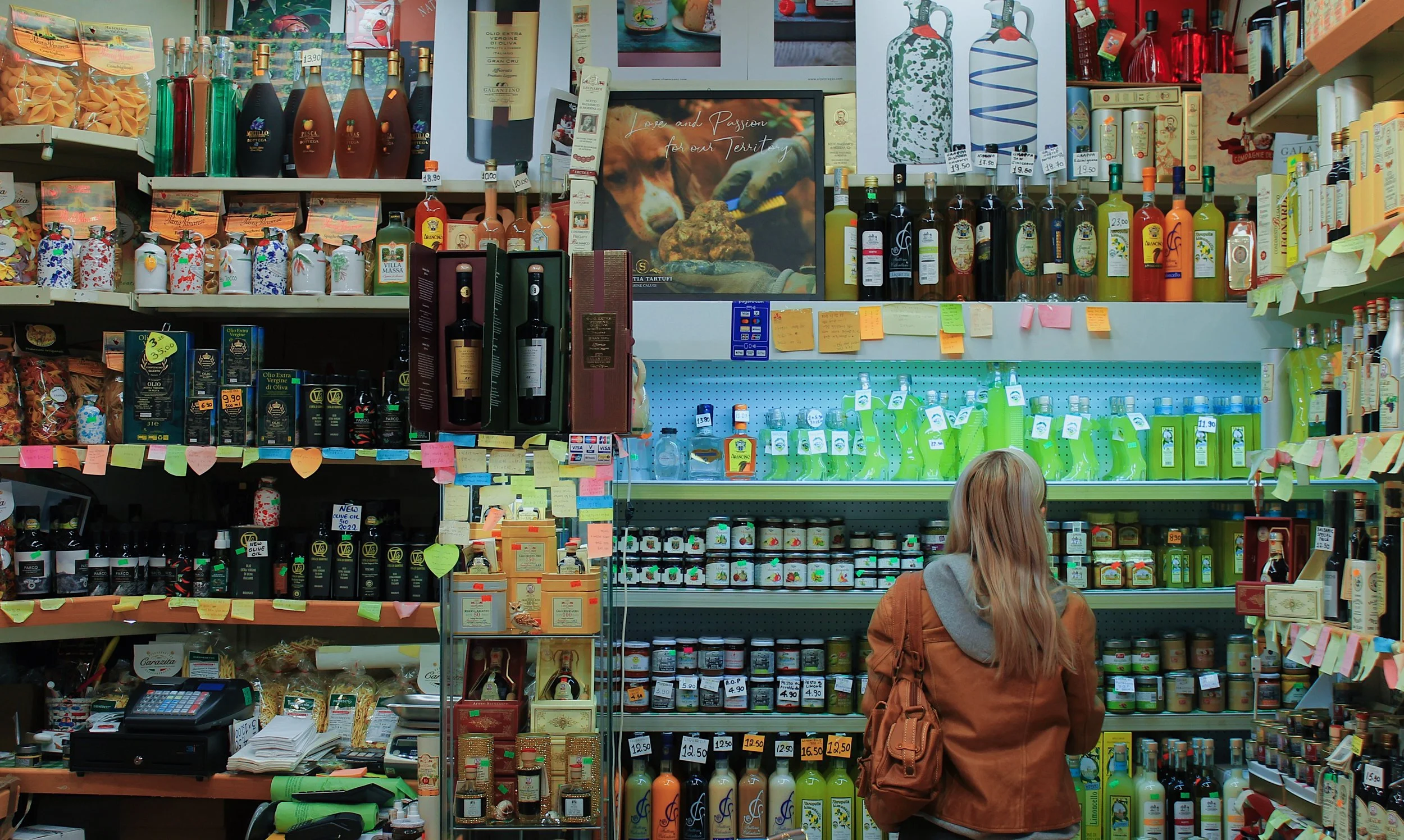 A woman shopping in Mercato Centrale in Florence, Italy. Filled with bottles of olive oils, condiments, limoncello, and packaged foods, with shelves full of colorful products and sticky notes attached to the shelves.
