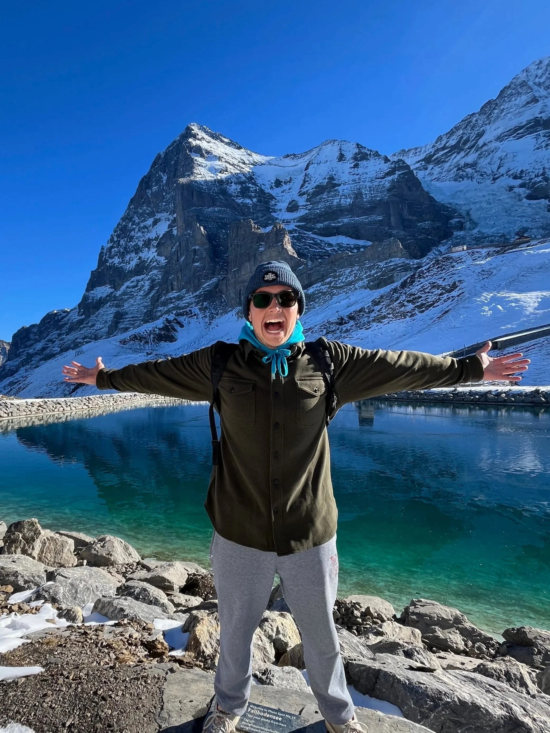 Person wearing sunglasses, a blue beanie, and outdoor clothing with arms outstretched on rocky terrain near a clear, reflective mountain lake with Swiss Alps in the background.