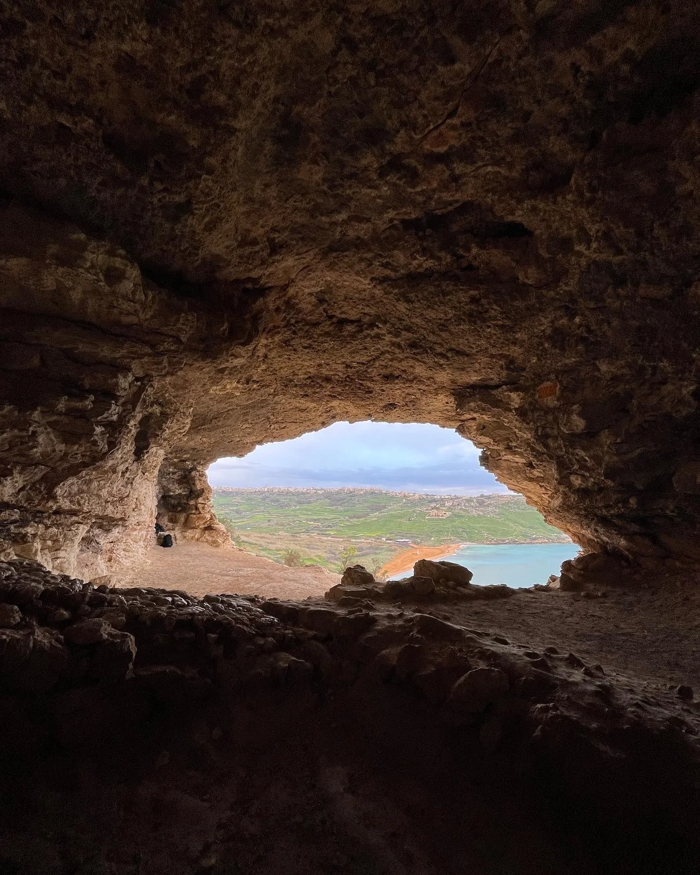 View through a natural cave opening in Malta, showing a coastal landscape with green fields, a body of water, and a distant city skyline under a cloudy sky.
