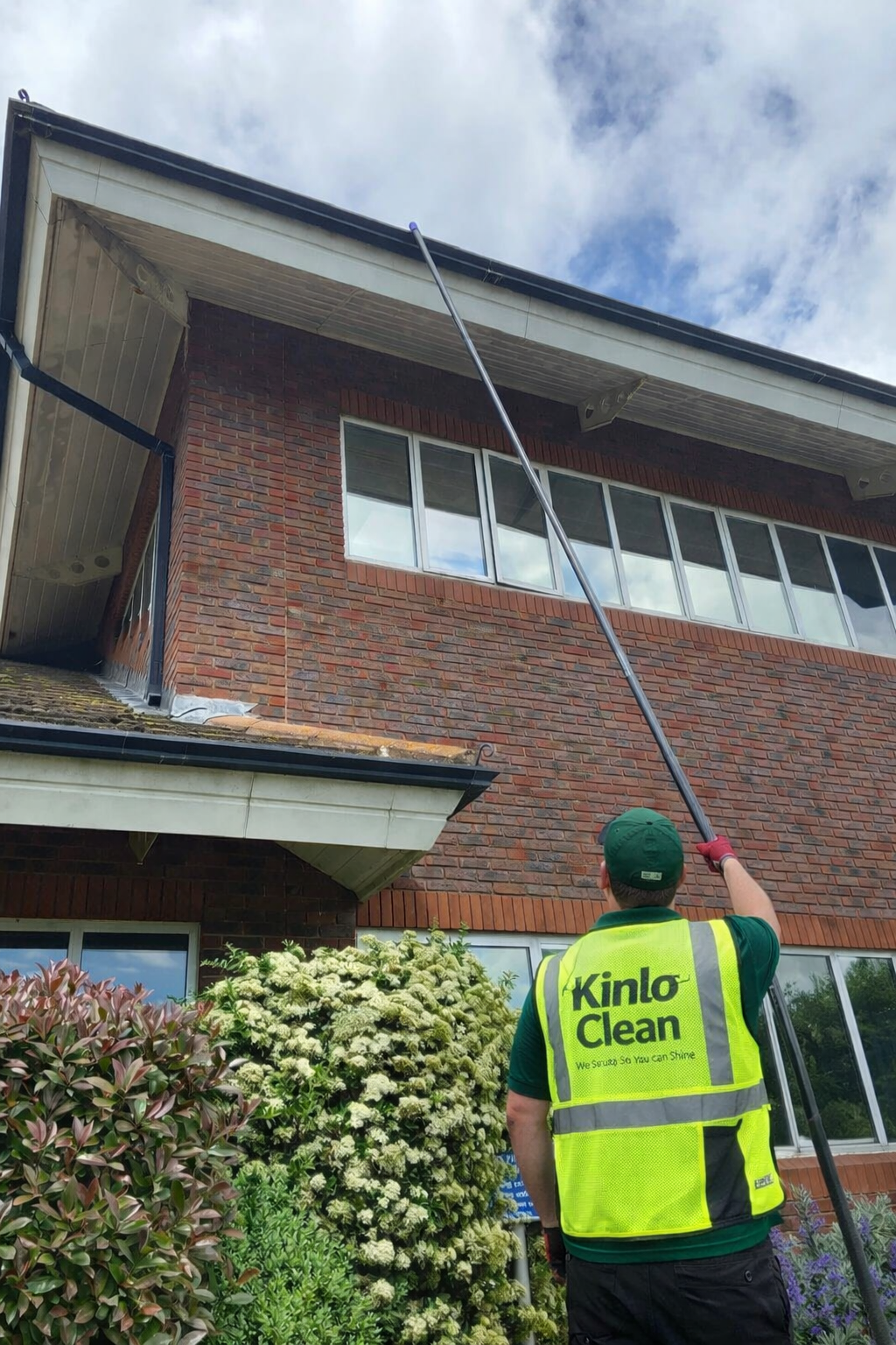A worker from Kinlo Clean using an extendable pole to clean the outside windows of a two-story brick building. The worker is wearing a high-visibility yellow vest with the company logo, green shirt, and a green cap. There are bushes with white flowers in the foreground, and the sky is partly cloudy.